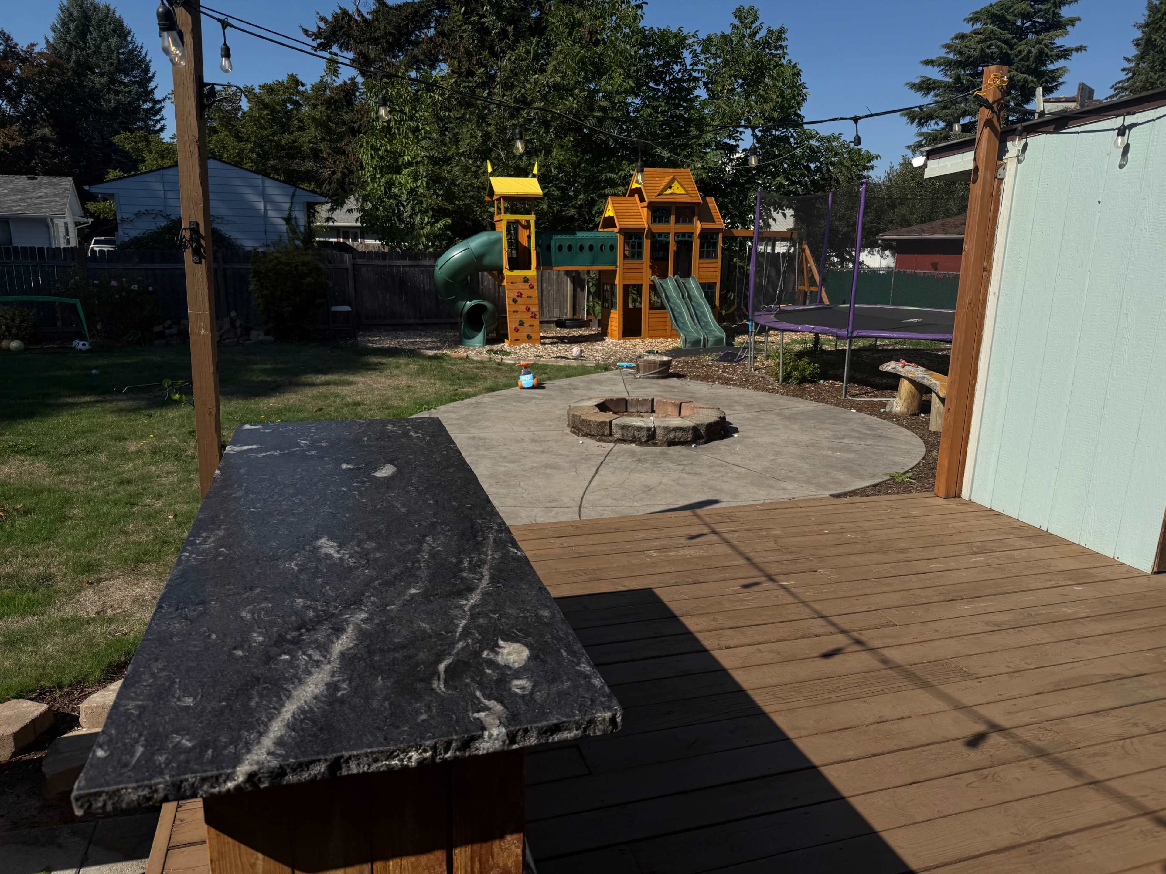 The image shows a backyard featuring a wooden deck with a stone countertop, a fire pit area, and a children's play structure with a slide and swings in the background.