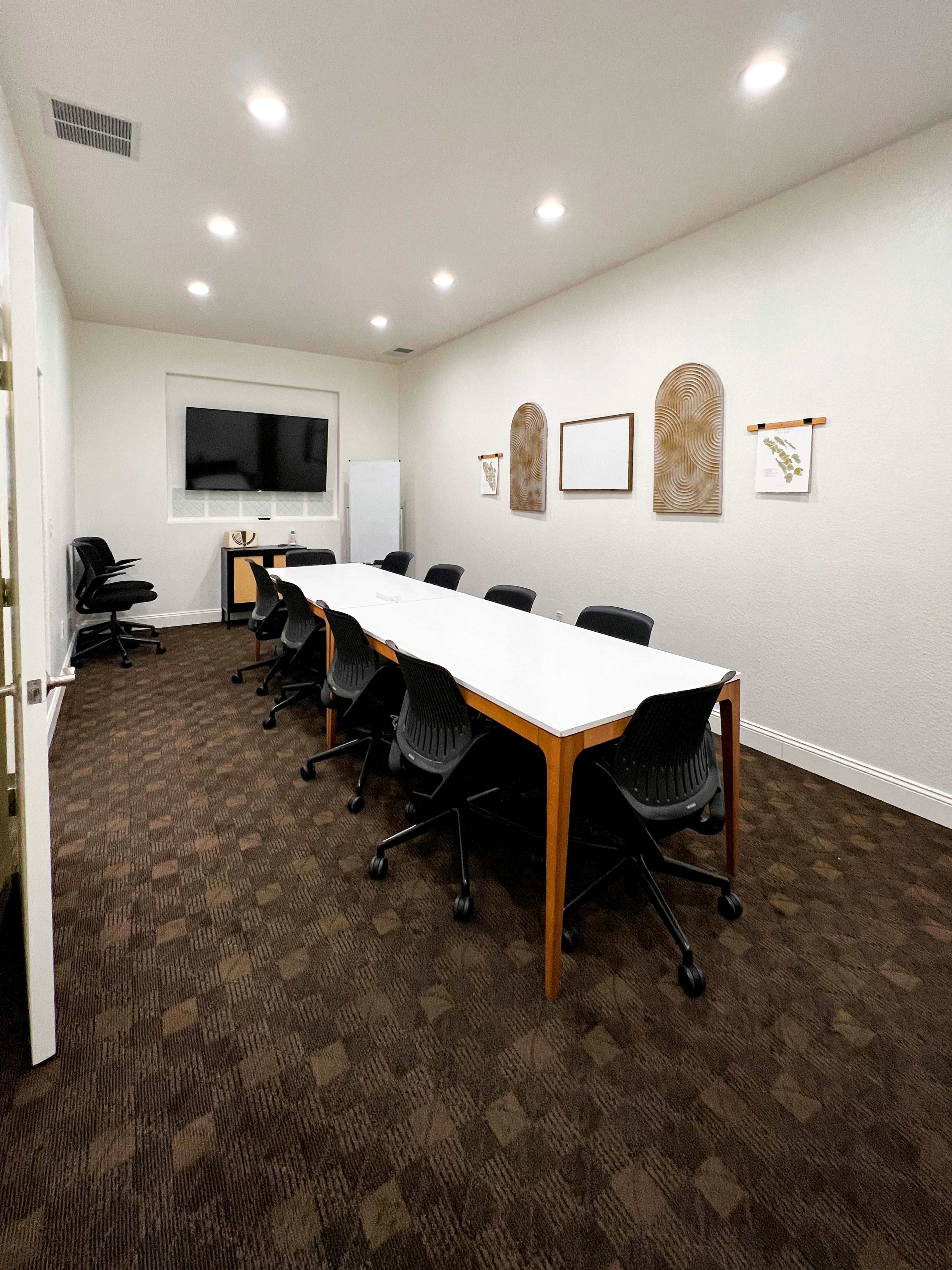 A modern conference room features a long white table with black chairs, illuminated by overhead lights and equipped with a wall-mounted TV.