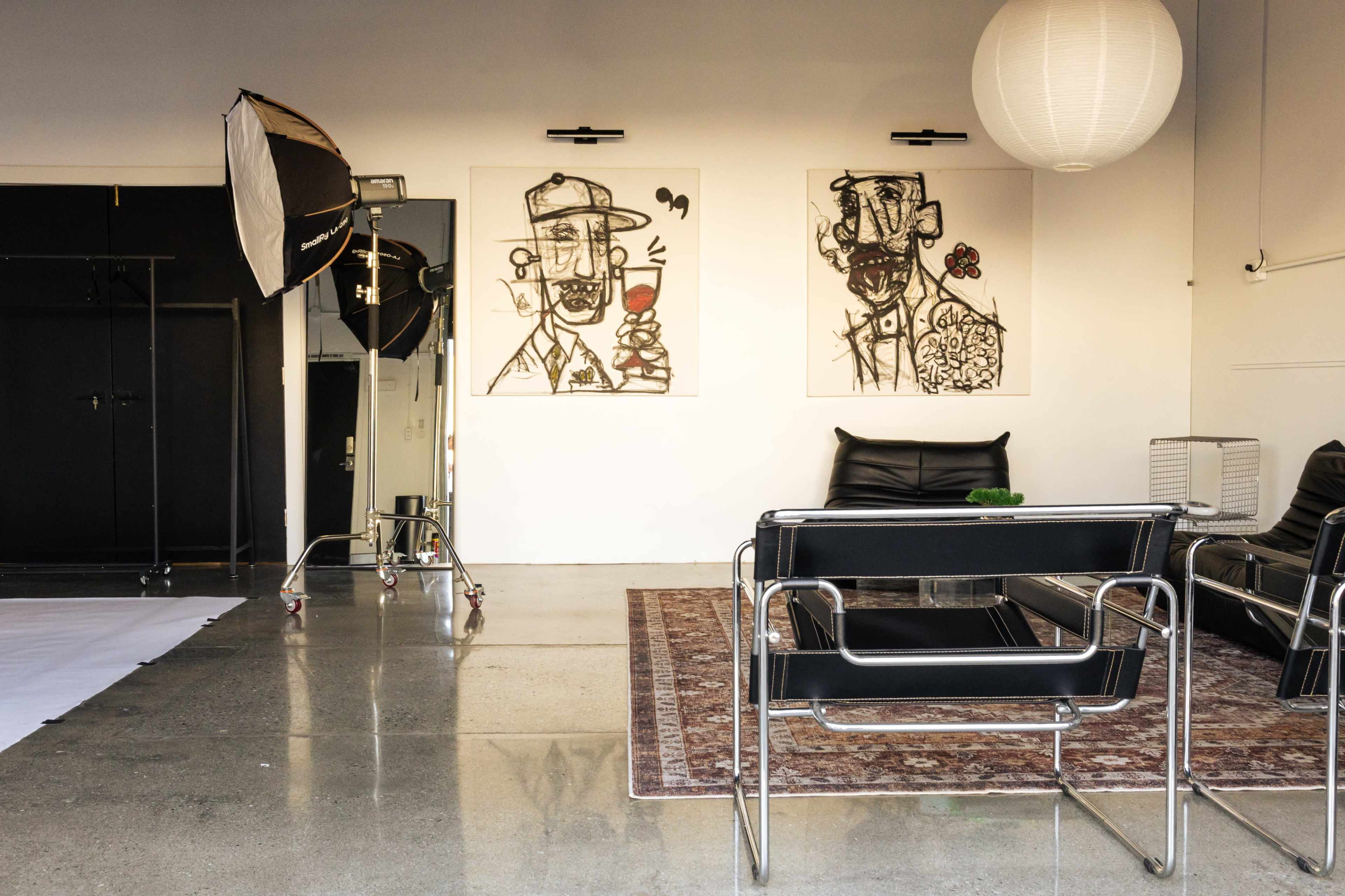 The image shows a modern interior space with two large framed artworks on the wall, a black leather sofa, a vintage rug, and studio lighting equipment.