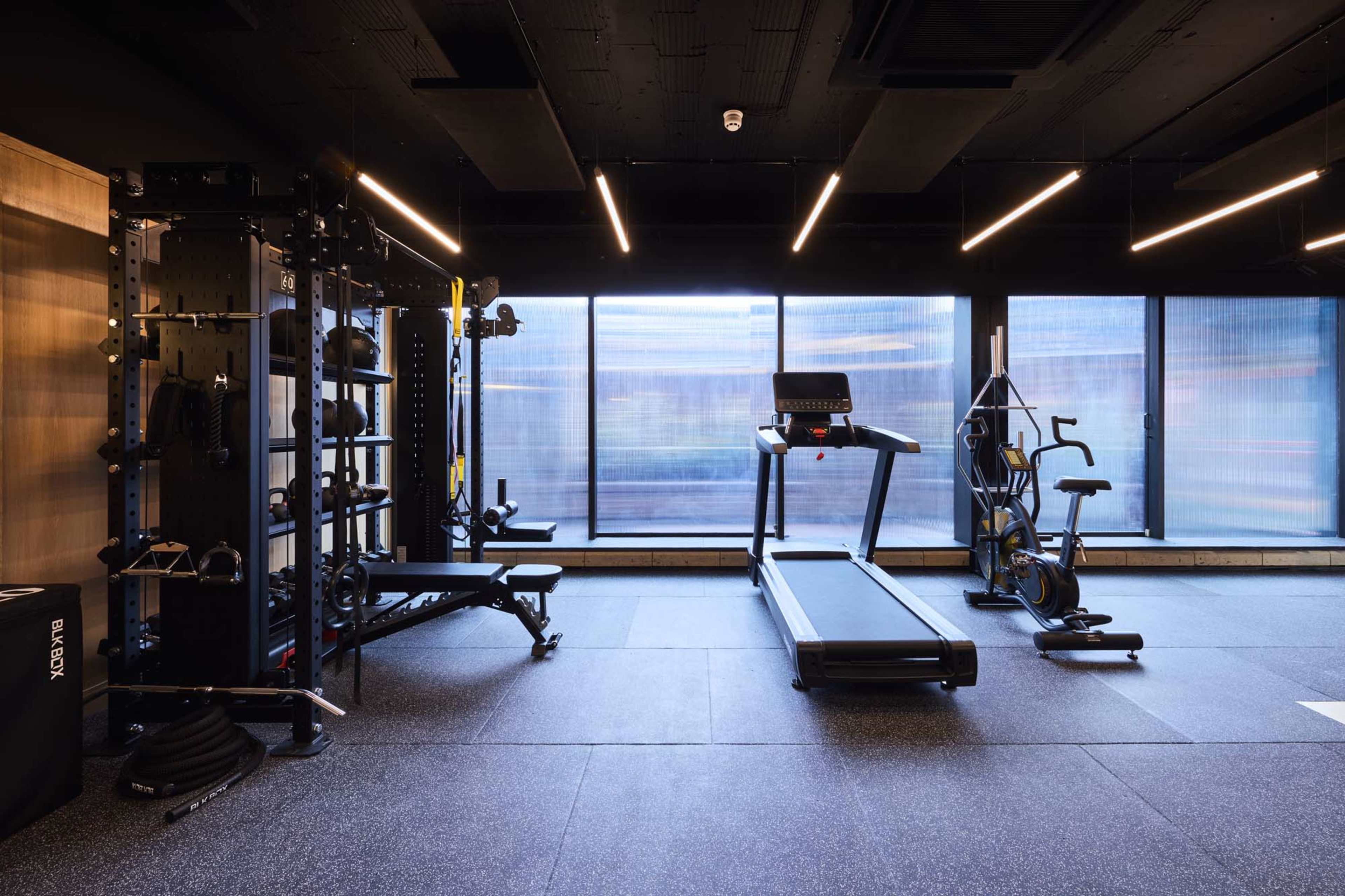 The image shows a modern gym featuring a treadmill, an exercise bike, a multi-station weight machine, and large windows allowing natural light to enter.