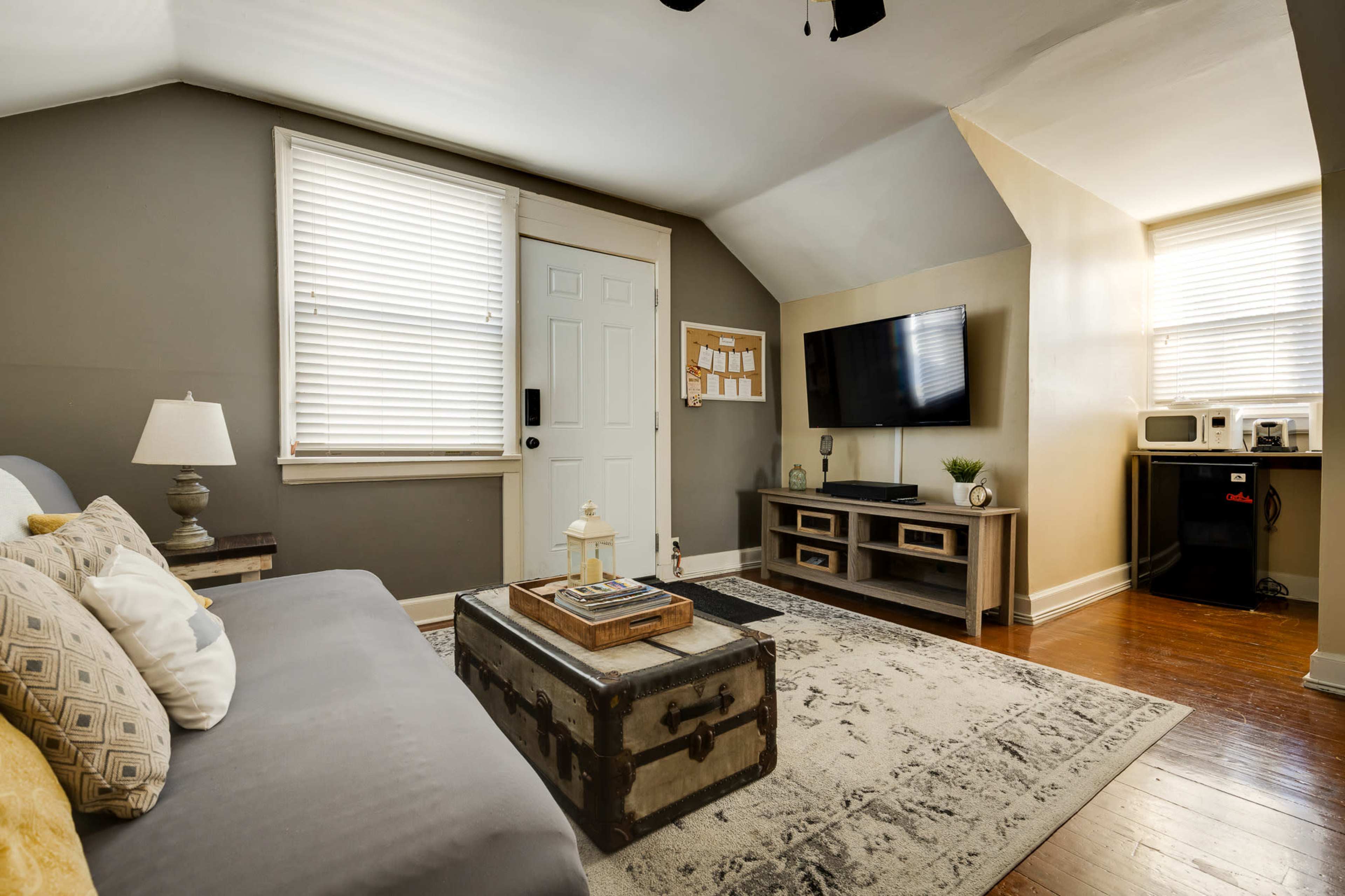 The image shows a cozy living room with a gray sofa, a wooden coffee table, a television mounted on the wall, and a small kitchenette area with a microwave and a refrigerator.