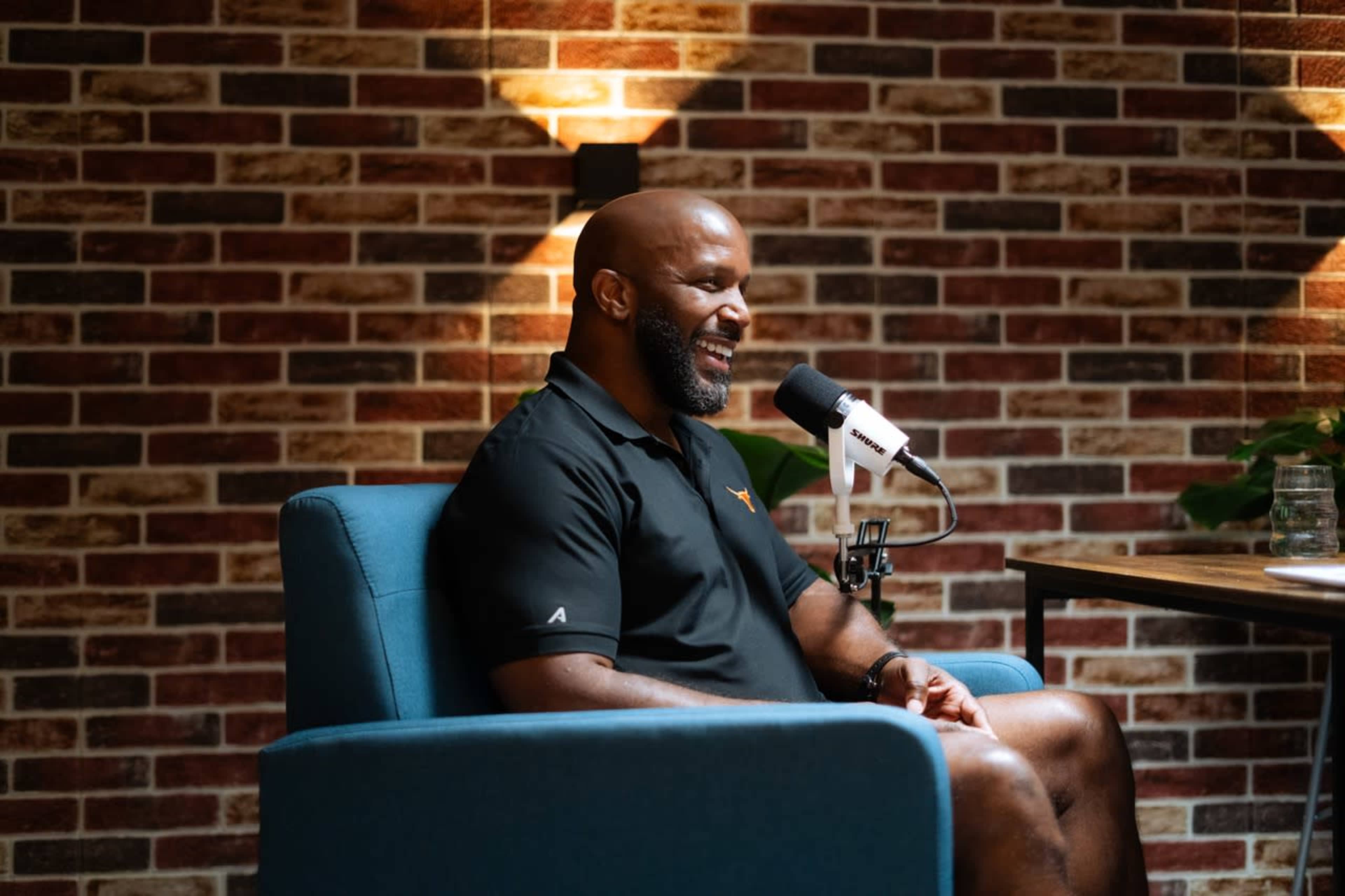 A man sits in a blue armchair with a microphone in front of him, smiling against a brick wall backdrop.