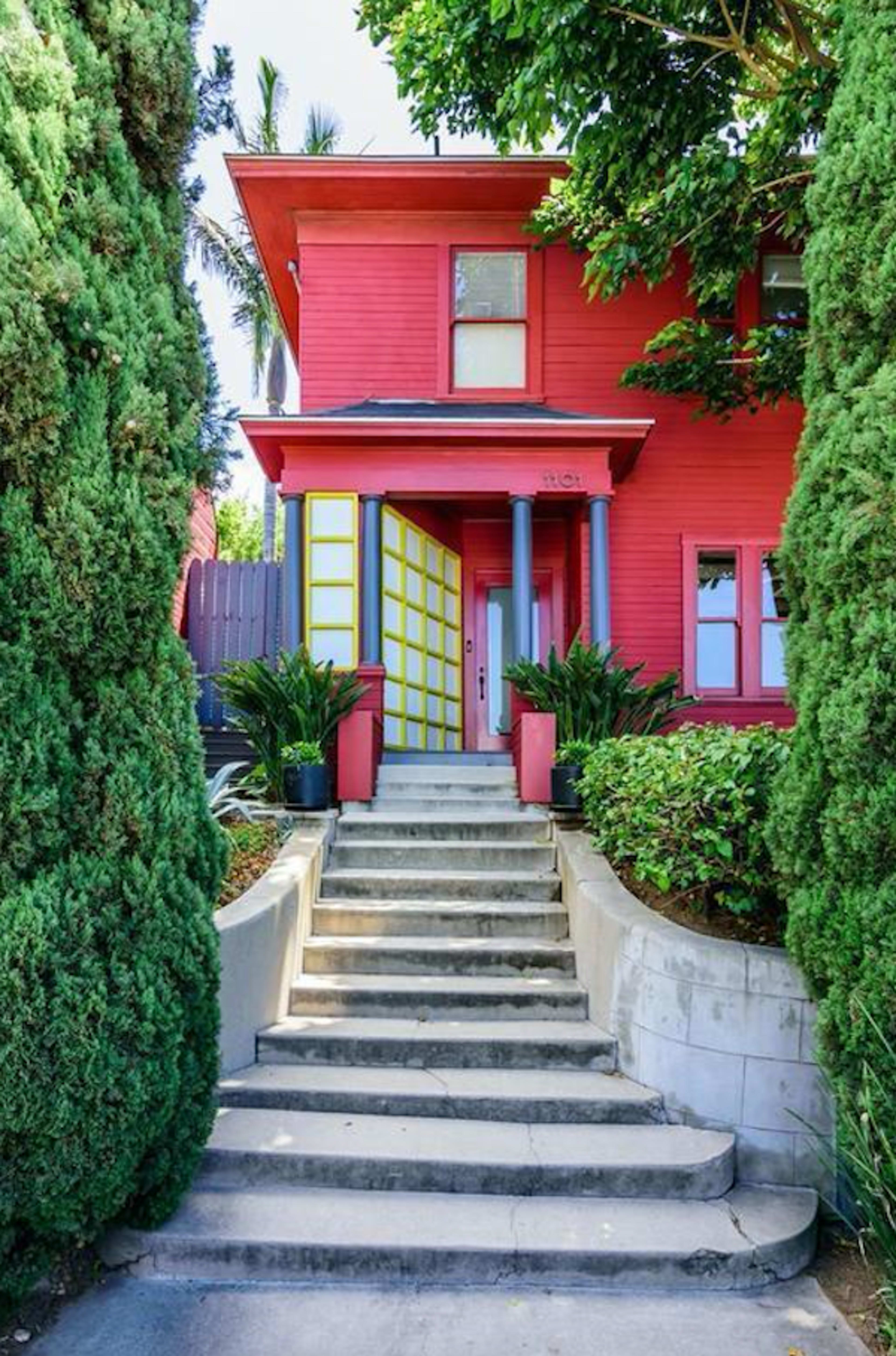 A bright red house with a yellow door is flanked by tall green shrubs and a stone staircase leading to the entrance.