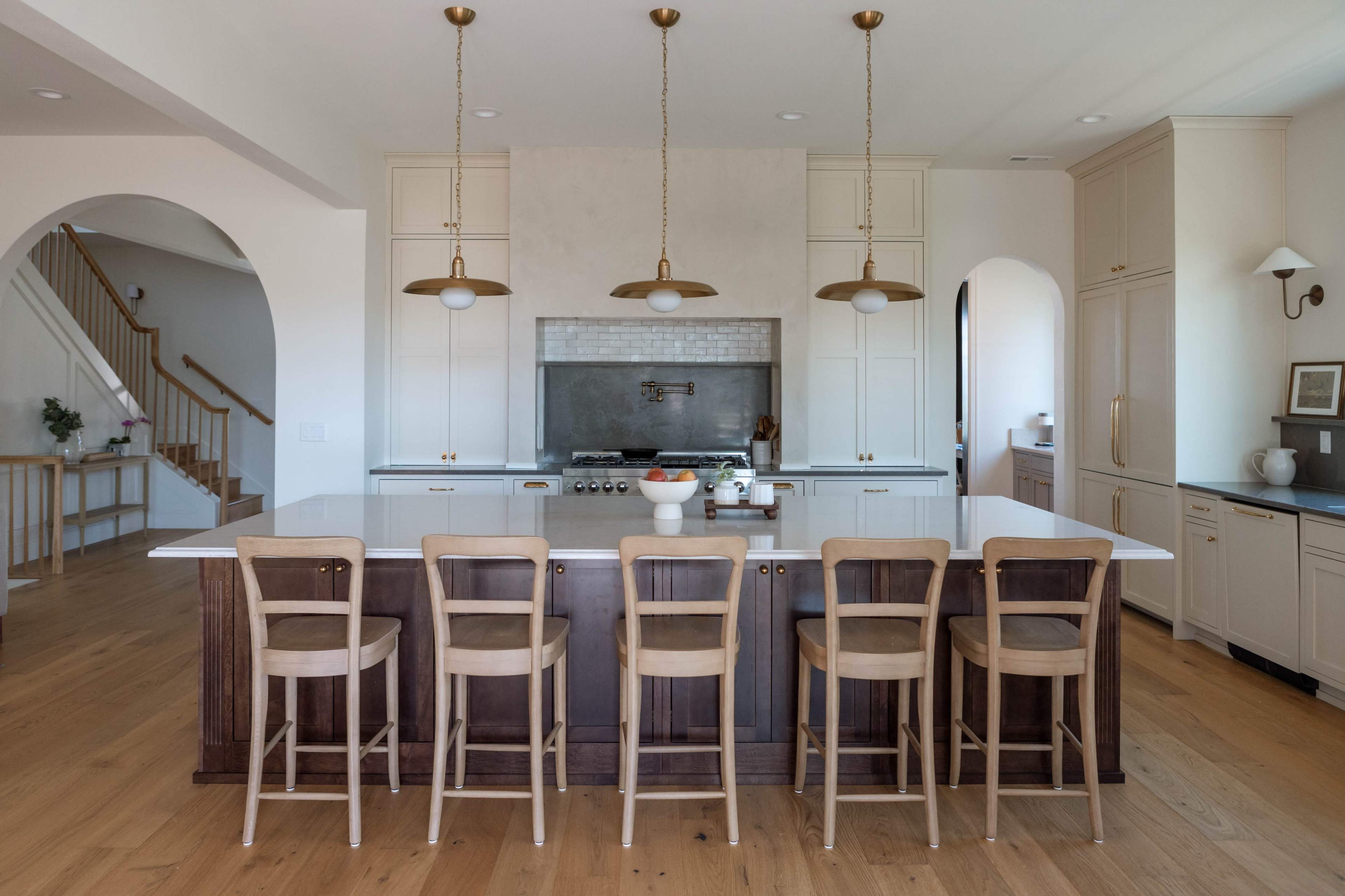 The image shows a modern kitchen with a large island, bar stools, and an arched entrance, featuring light-colored cabinetry and pendant lighting.