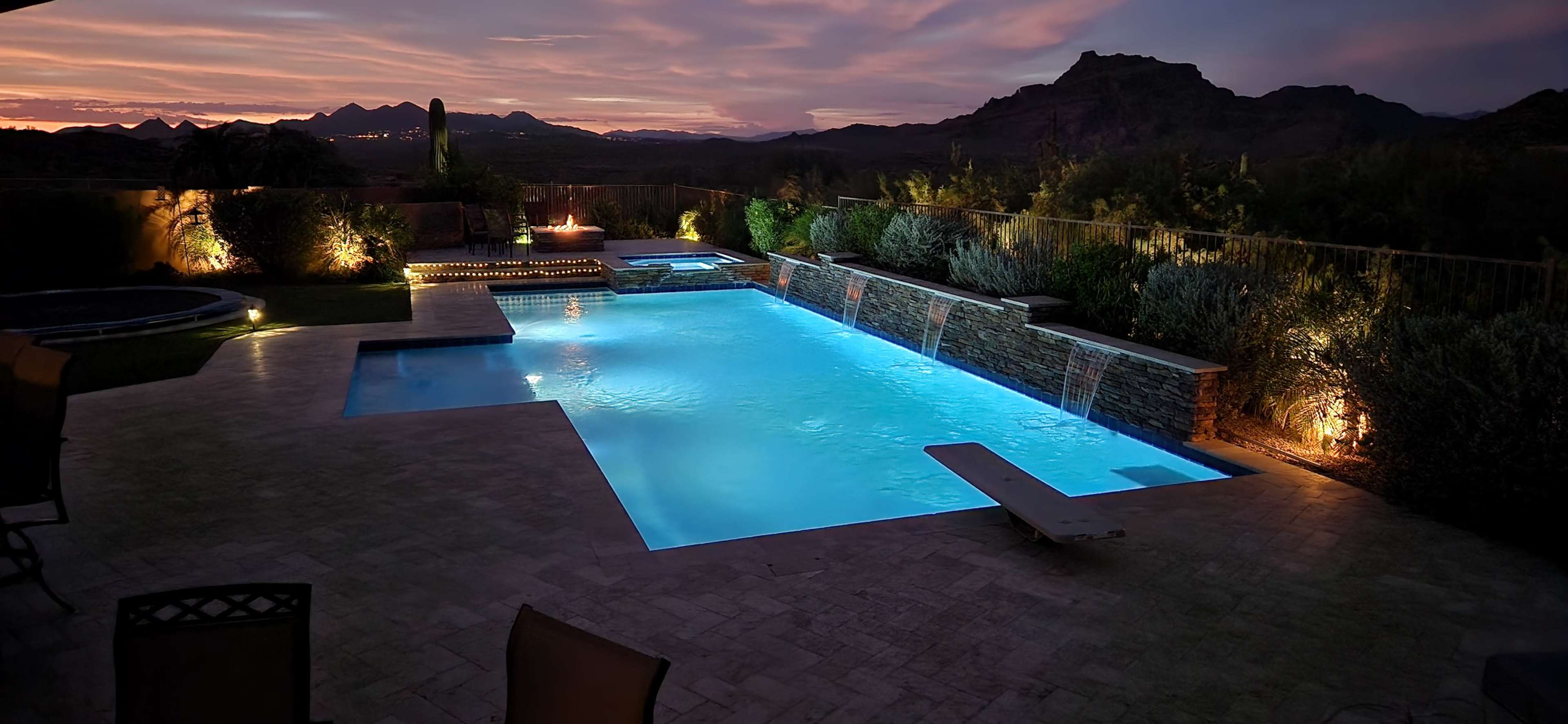 A lit swimming pool with water features and a diving board, surrounded by landscaped gardens and mountains under a twilight sky.