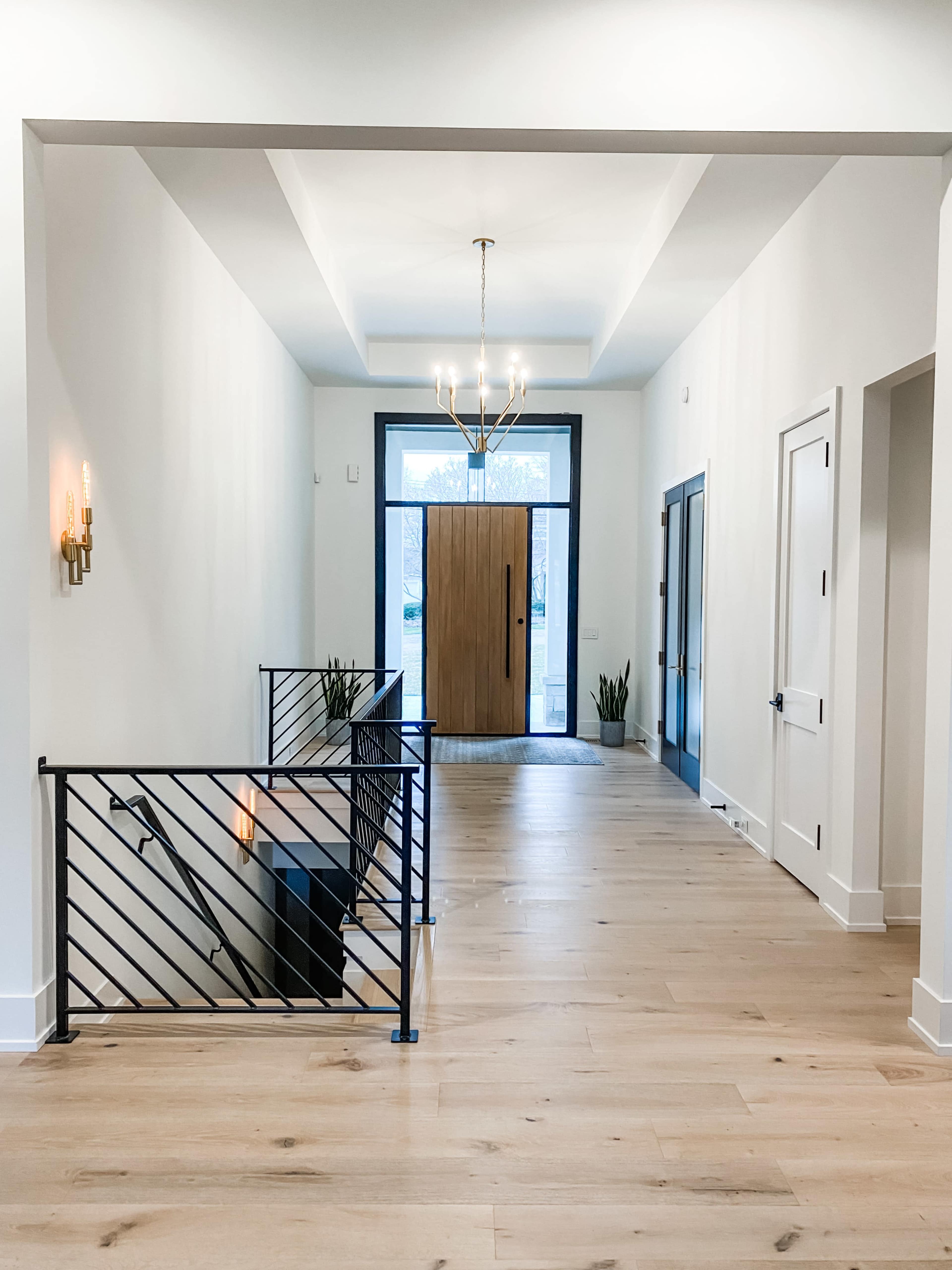 A spacious hallway features a modern staircase with black railings, a large front door at the end, and minimalist decor with hardwood flooring.