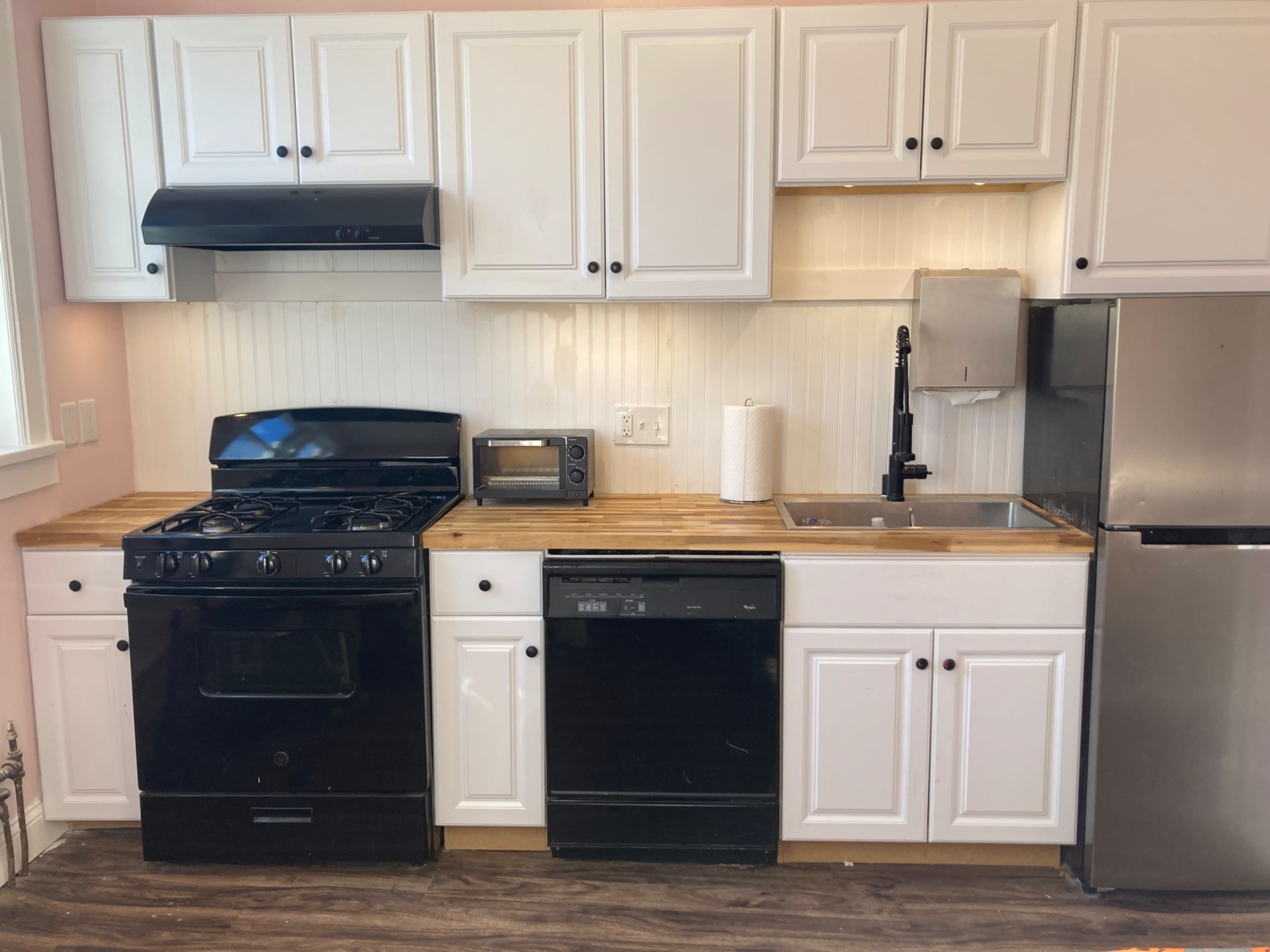 The image shows a kitchen with white cabinetry, a gas stove, a microwave, a sink, a dishwasher, and a stainless steel refrigerator along a wooden countertop.