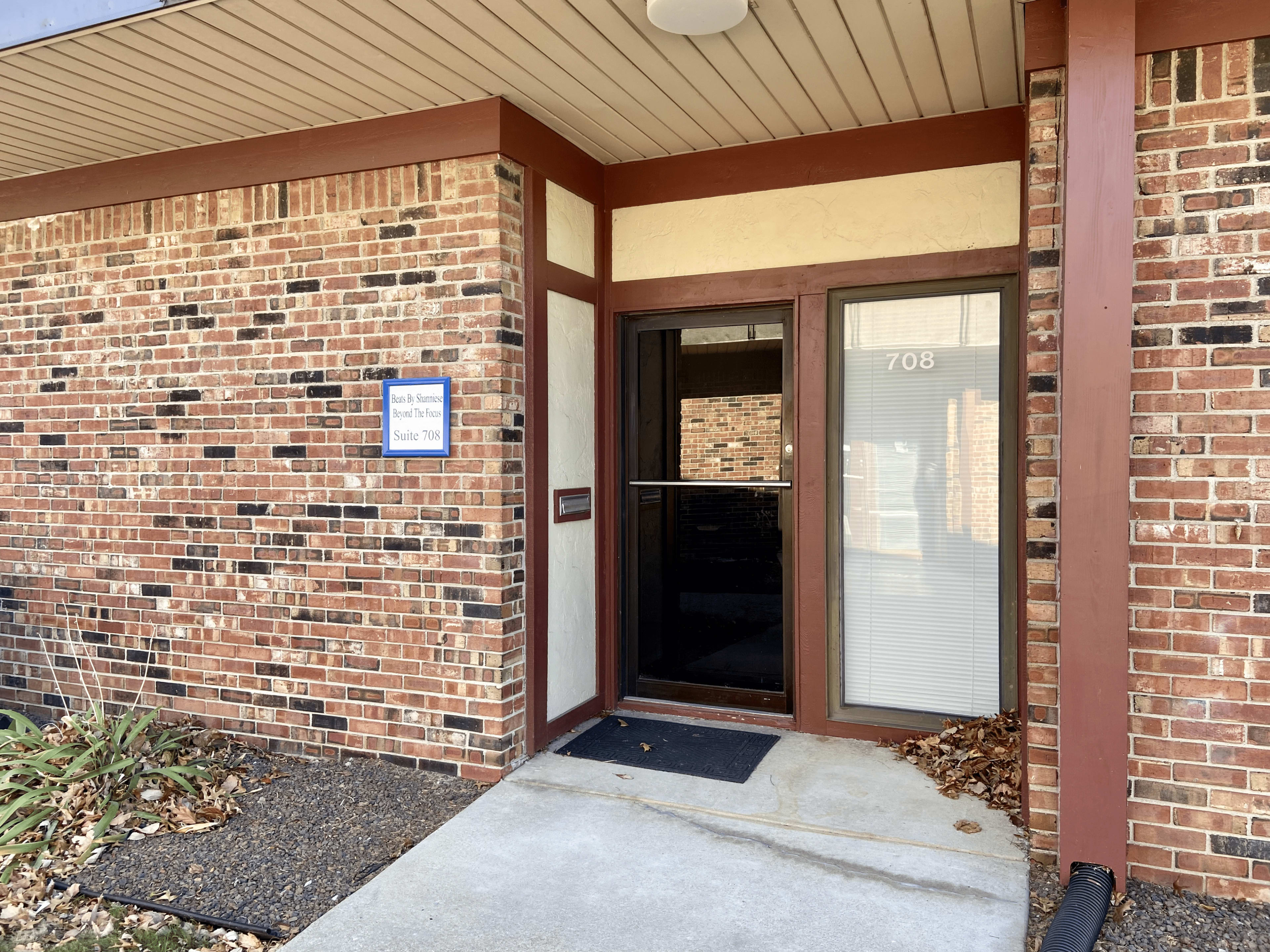 The entrance of a brick building features a glass door with a number 708 displayed next to it, alongside a sign mounted on the wall.