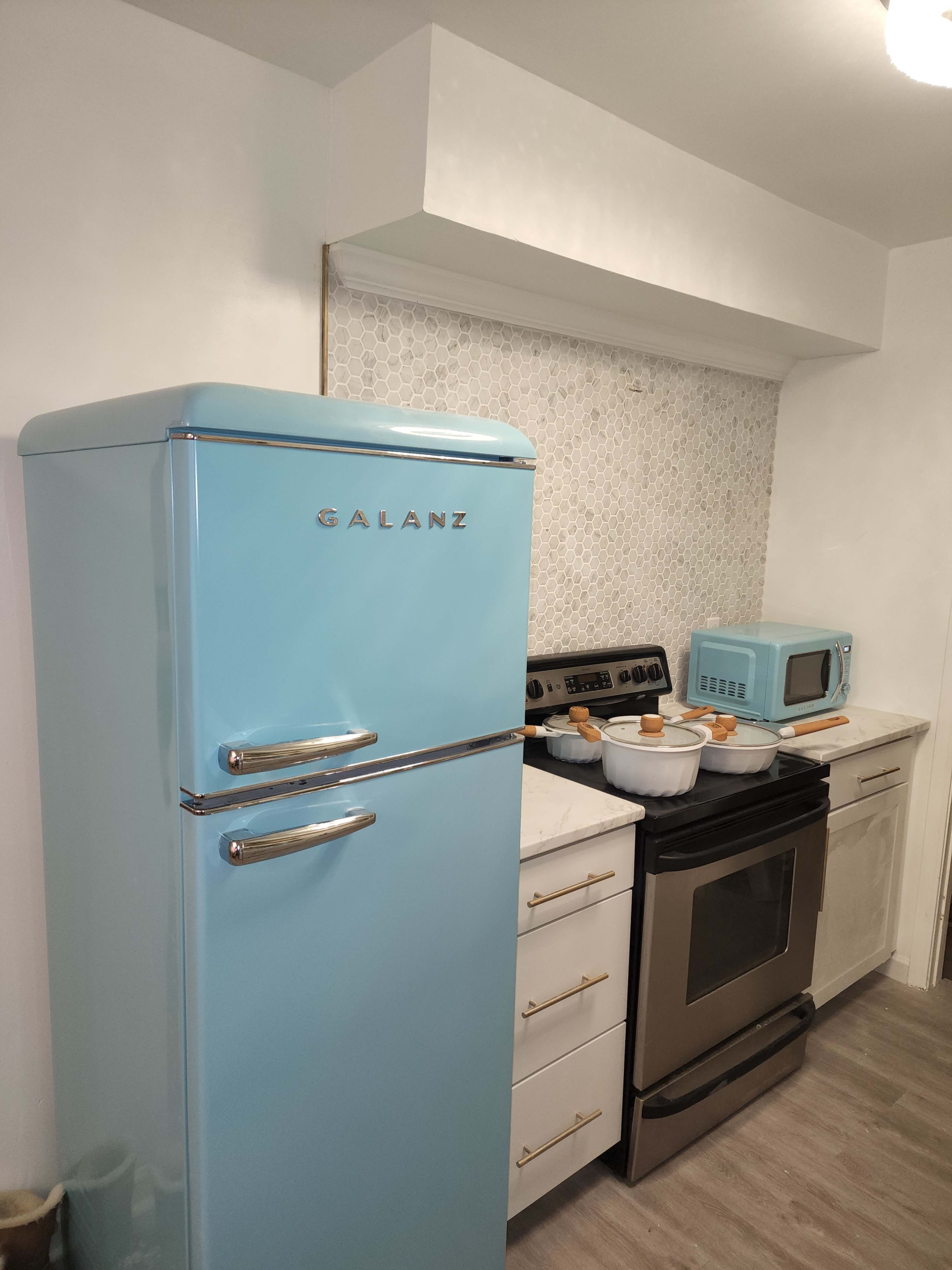 A kitchen featuring a retro-style light blue refrigerator beside an oven and a matching microwave, with a tiled backsplash and white cabinetry.