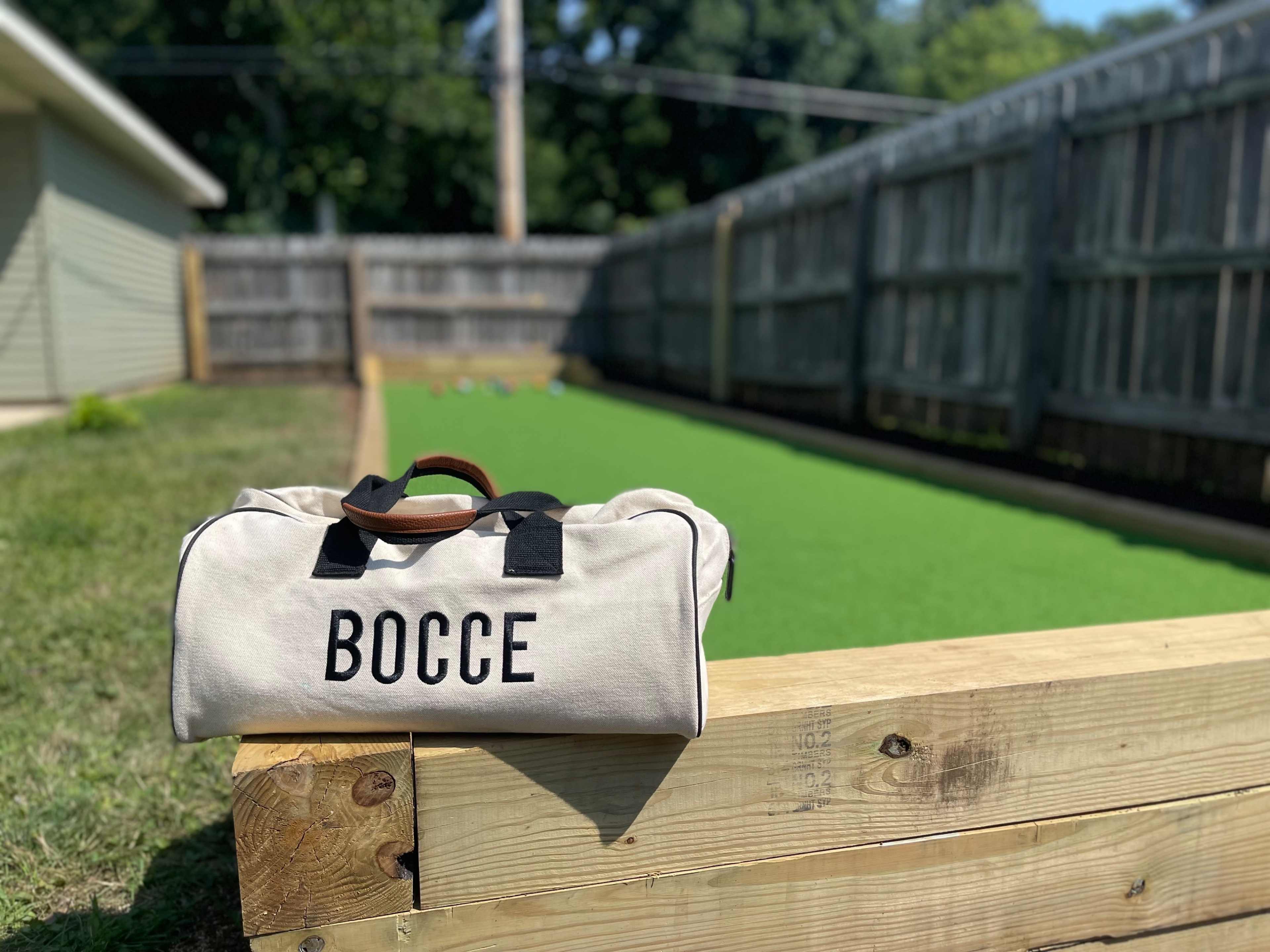 A canvas bag labeled "BOCCE" rests on a wooden ledge near a bocce ball court covered in green turf, surrounded by wooden fencing.