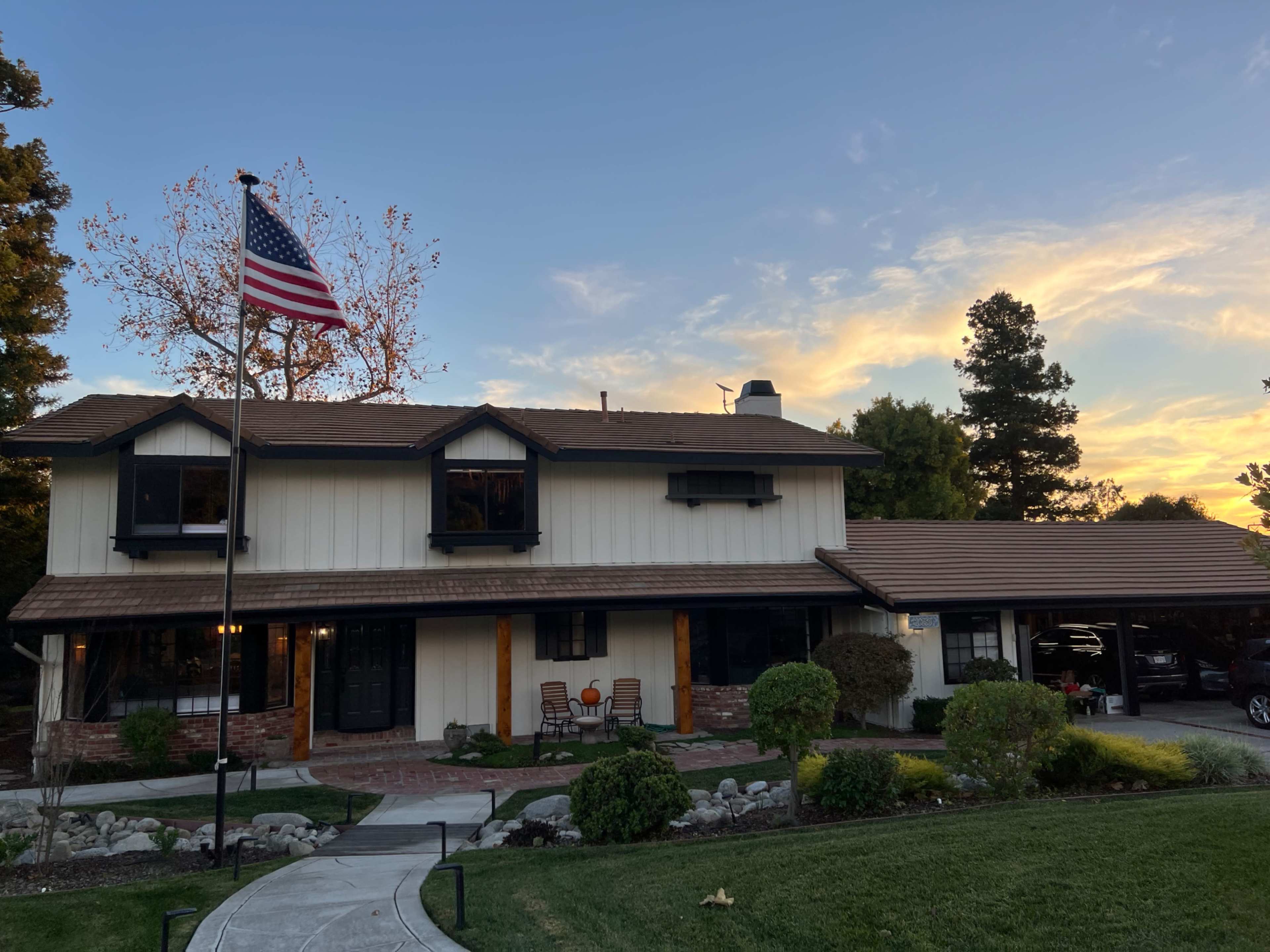 The image shows a two-story house with a flagpole displaying the American flag, surrounded by a lawn and landscaped garden, under a sunset sky.