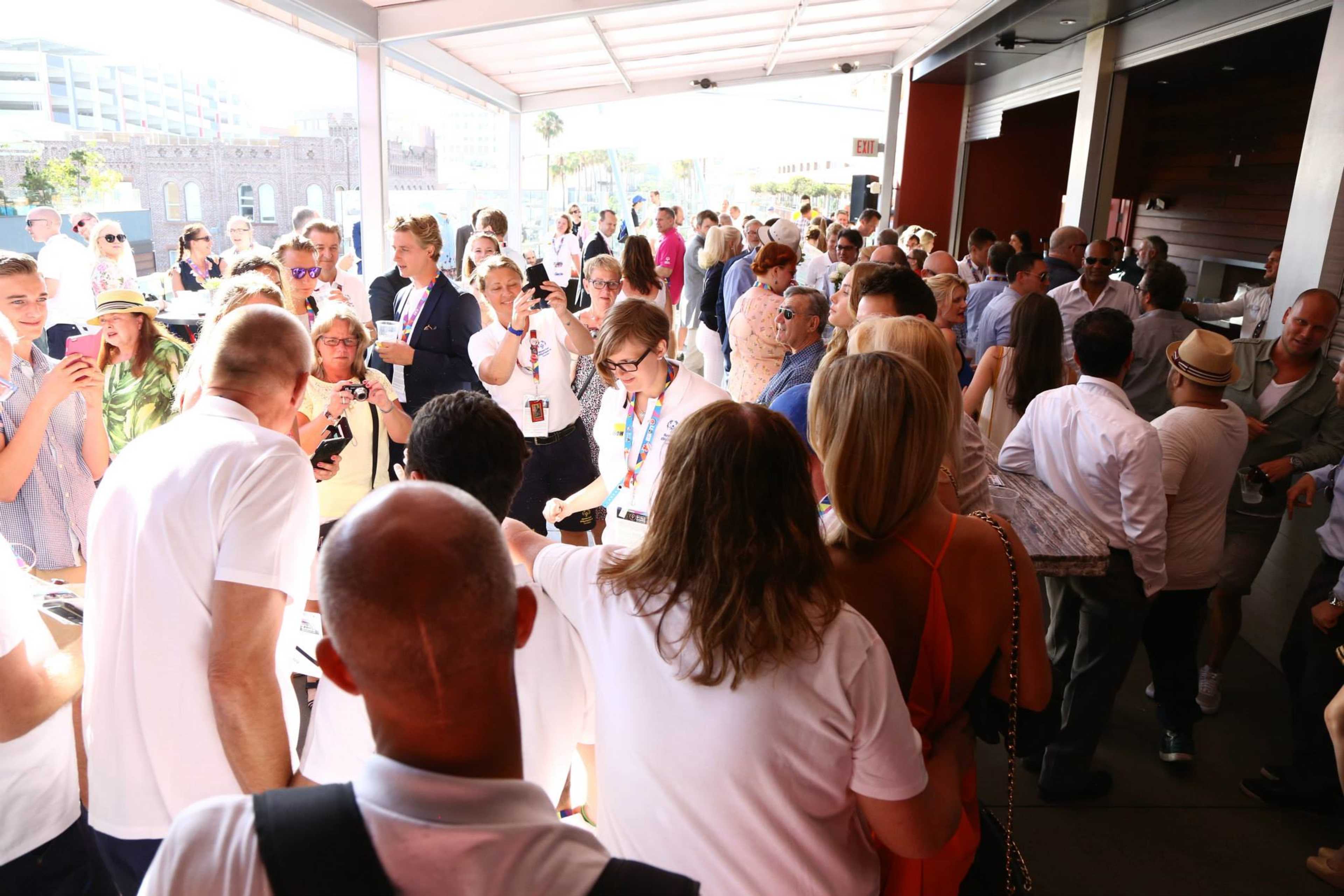 A crowded outdoor event features a mix of people engaging in conversation and using smartphone cameras.