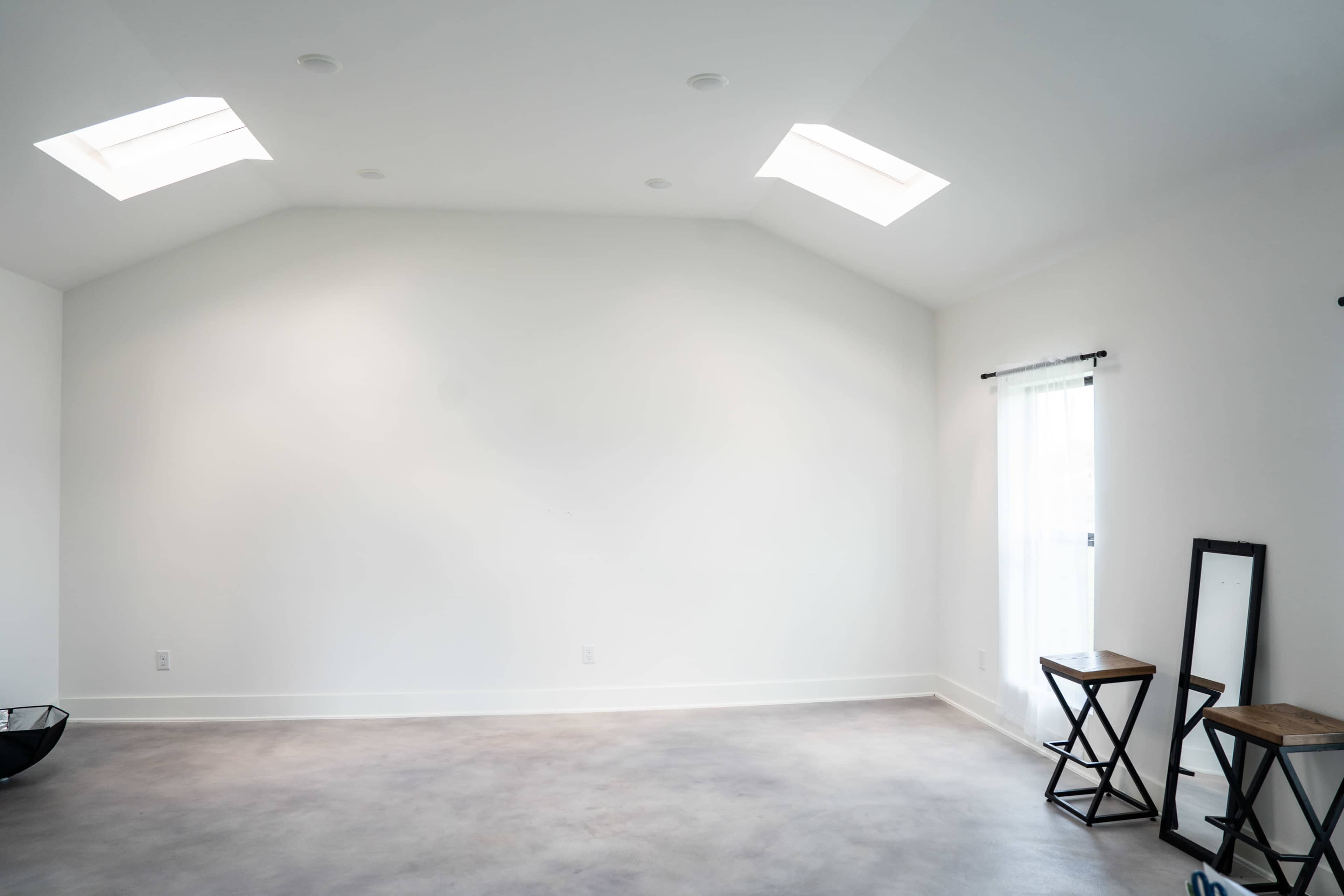 The image shows a spacious room with a high, white ceiling, two skylights, and minimal furnishings, including two wooden stools and a floor mirror.