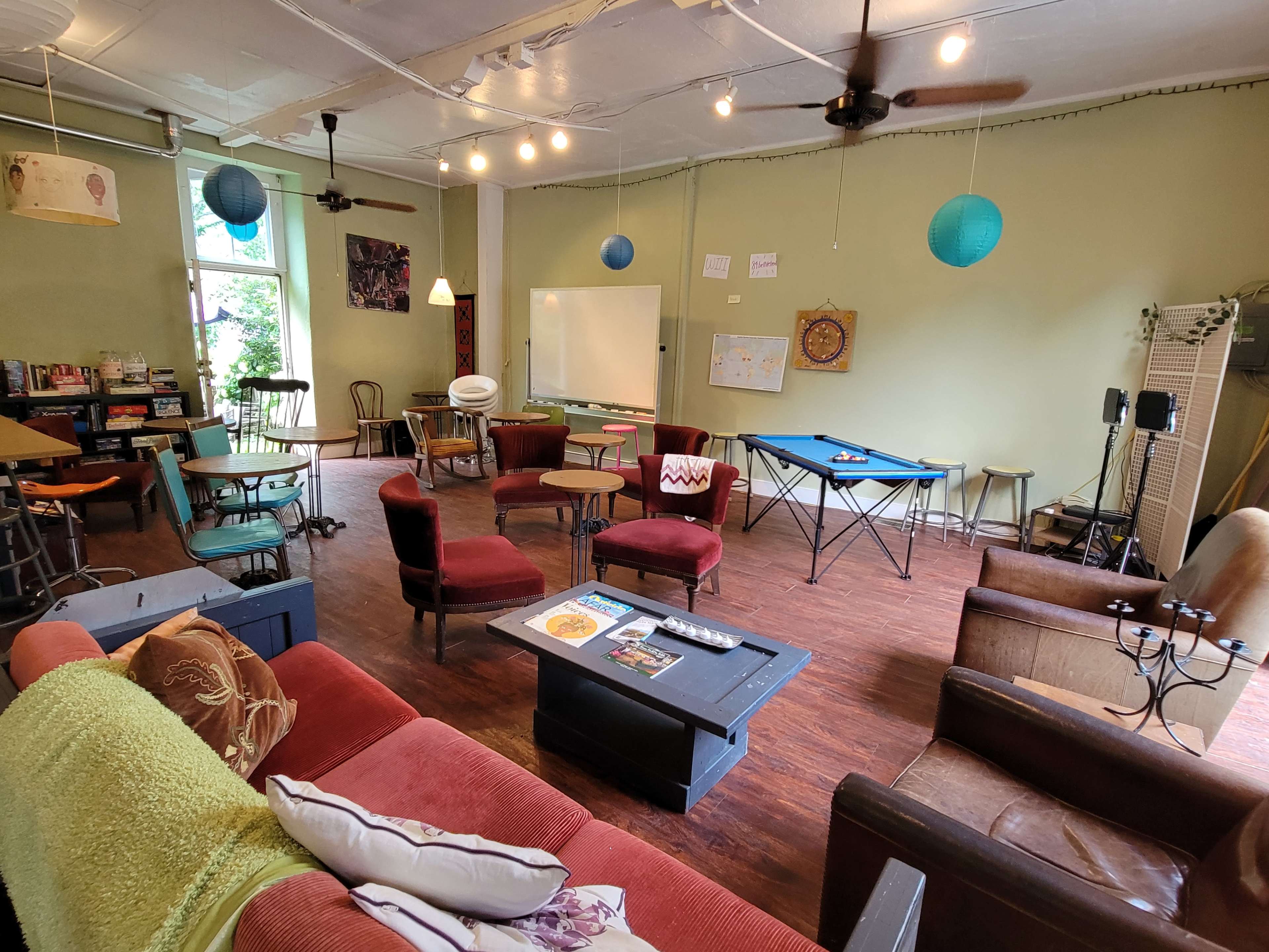 The image shows a cozy multi-purpose room furnished with seating, a table, and decorative elements, featuring green walls and ceiling lights.