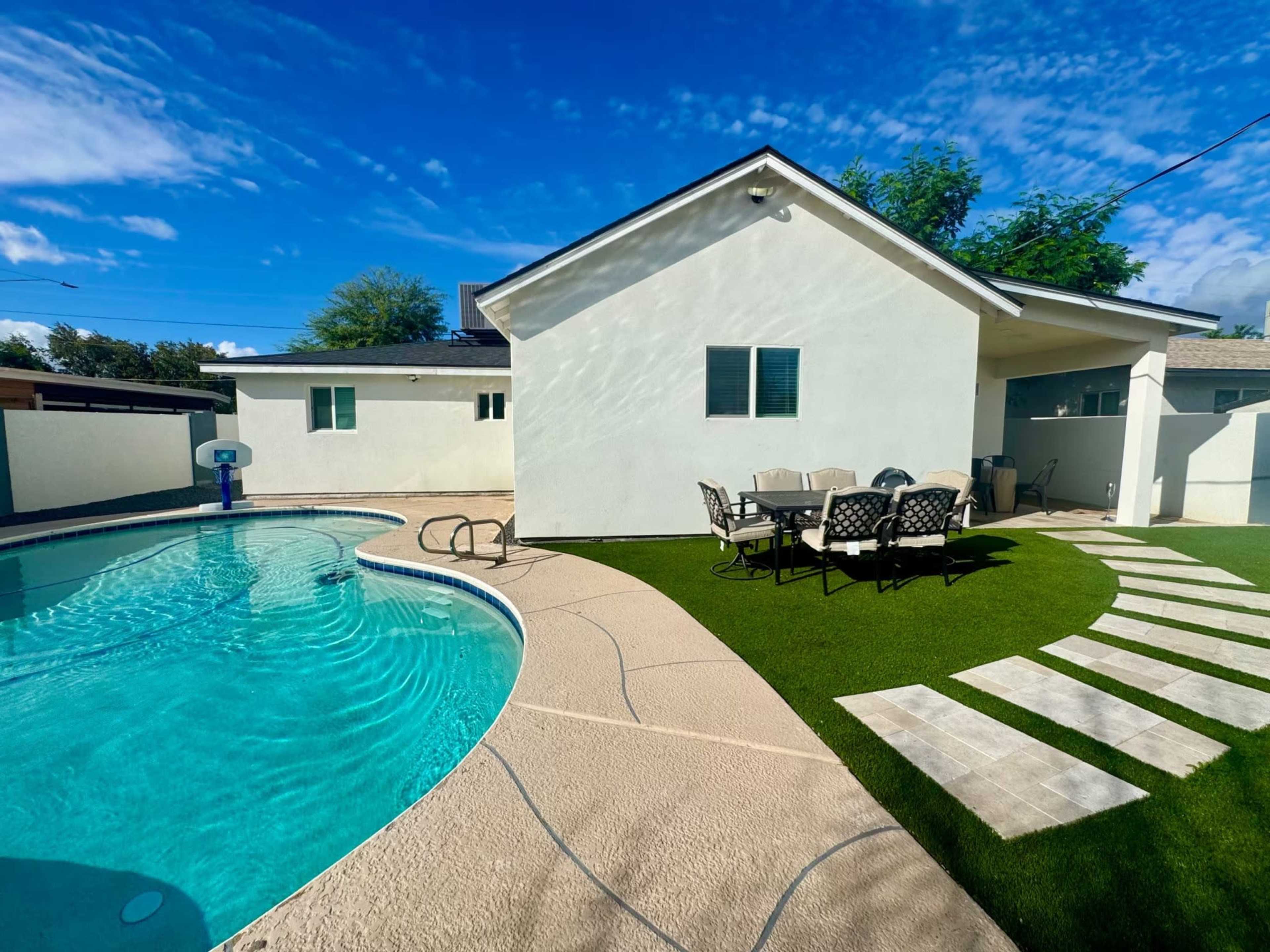 The image shows a backyard with a swimming pool, a patio area with a dining table, and a pathway leading to a white house under a clear blue sky.