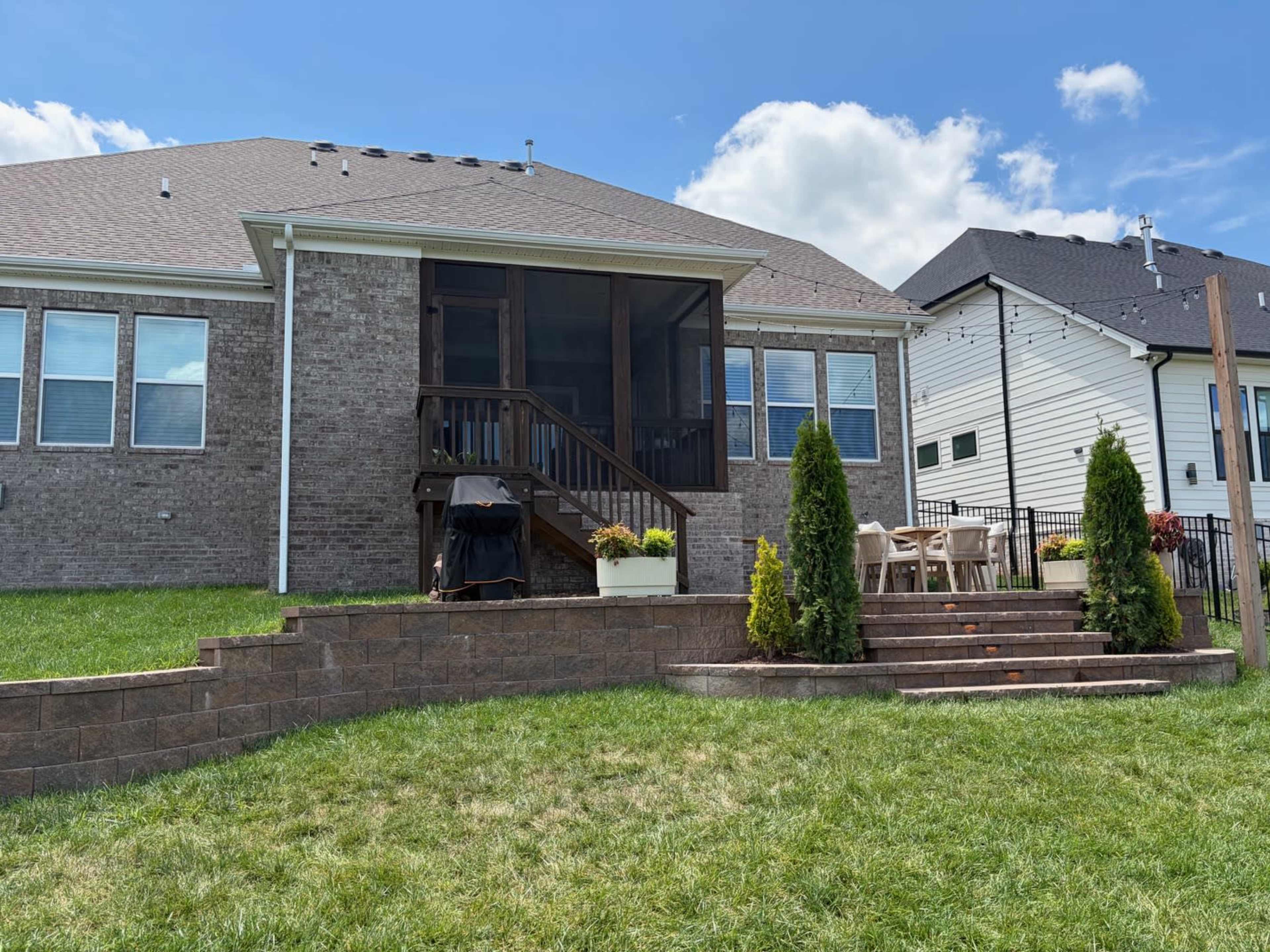 A brick house features a screened porch, a black grill, and a tiered garden with potted plants and seating areas.