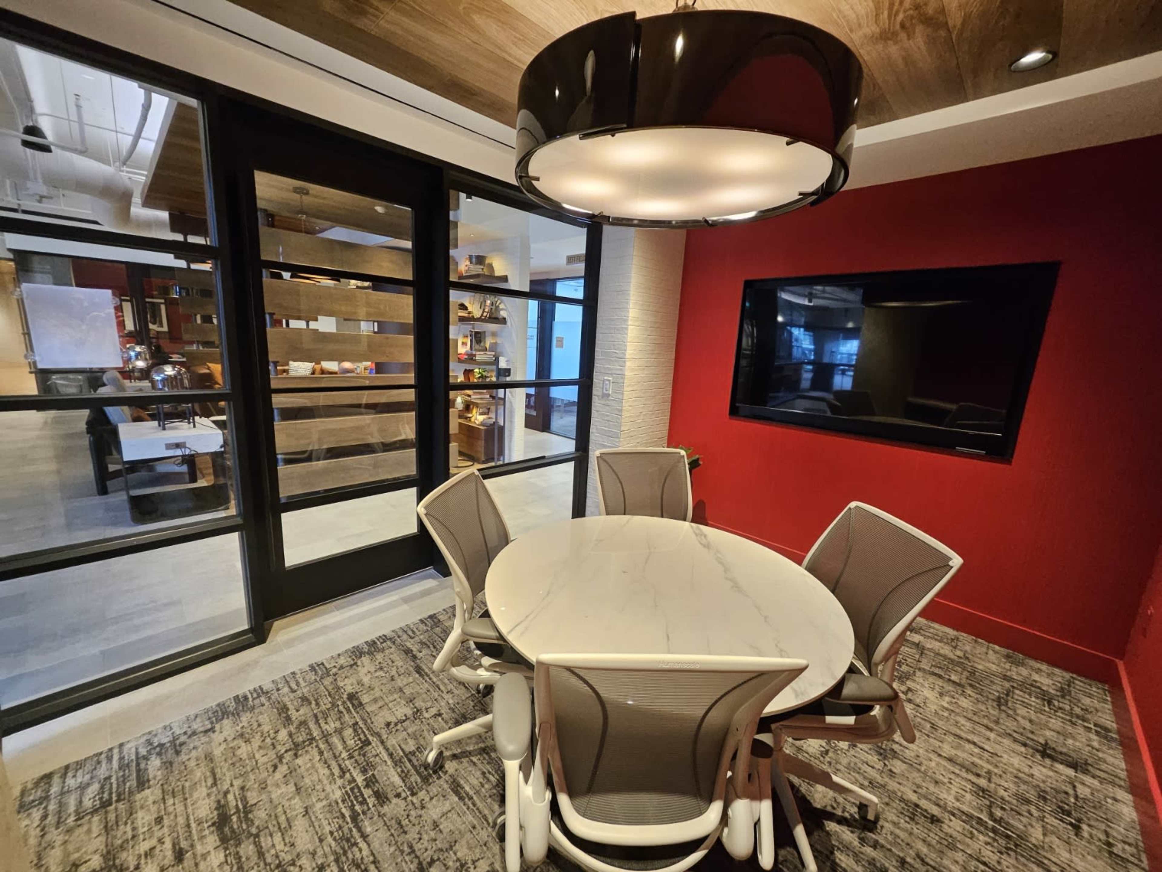 A conference room features a circular marble table surrounded by four chairs, a large overhead light, and a wall-mounted television, with glass partitions revealing a modern office space beyond.