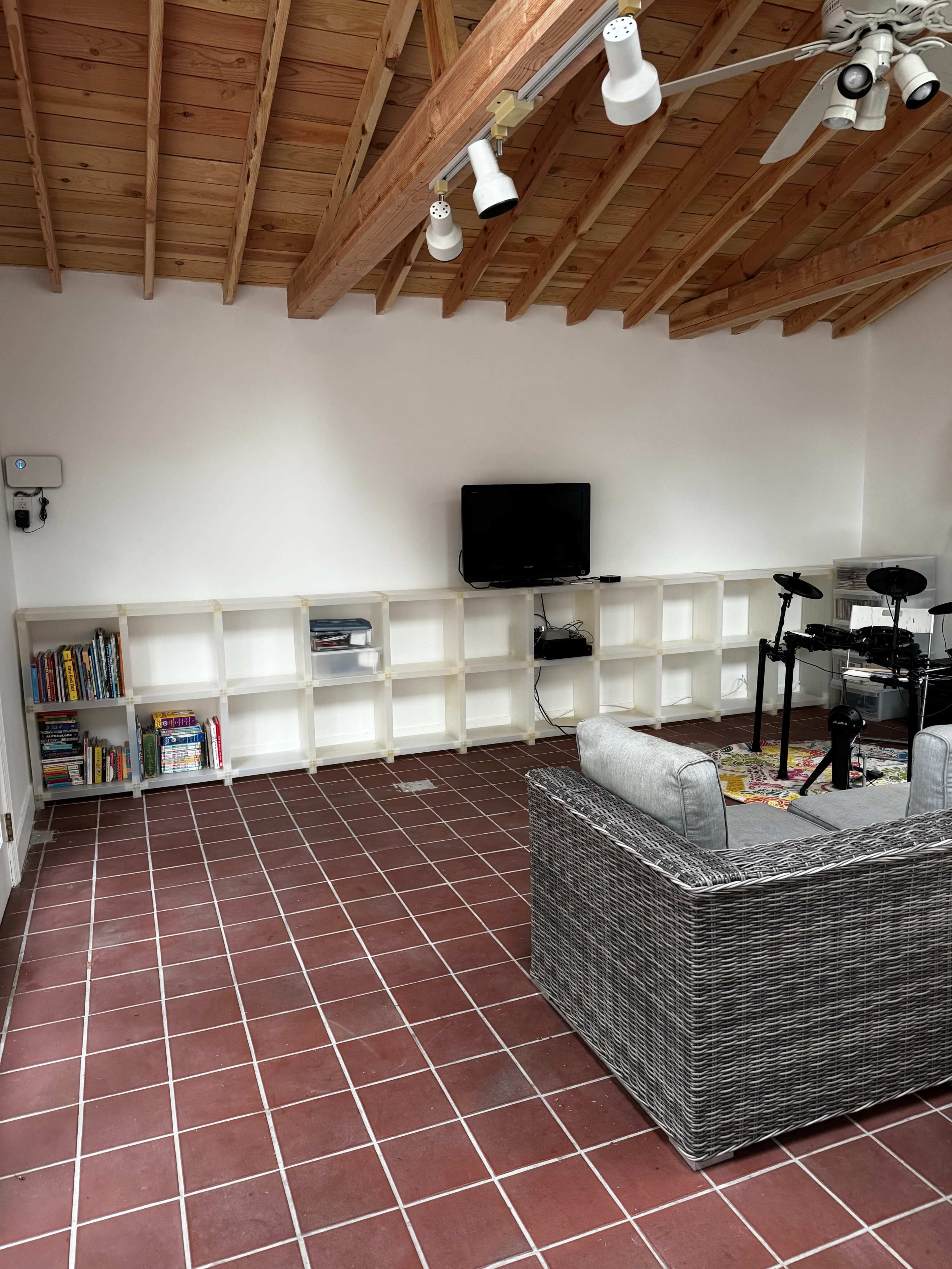 The image shows a spacious room with a tiled floor, a wall-mounted television, a gray sofa, and a bookcase filled with books.