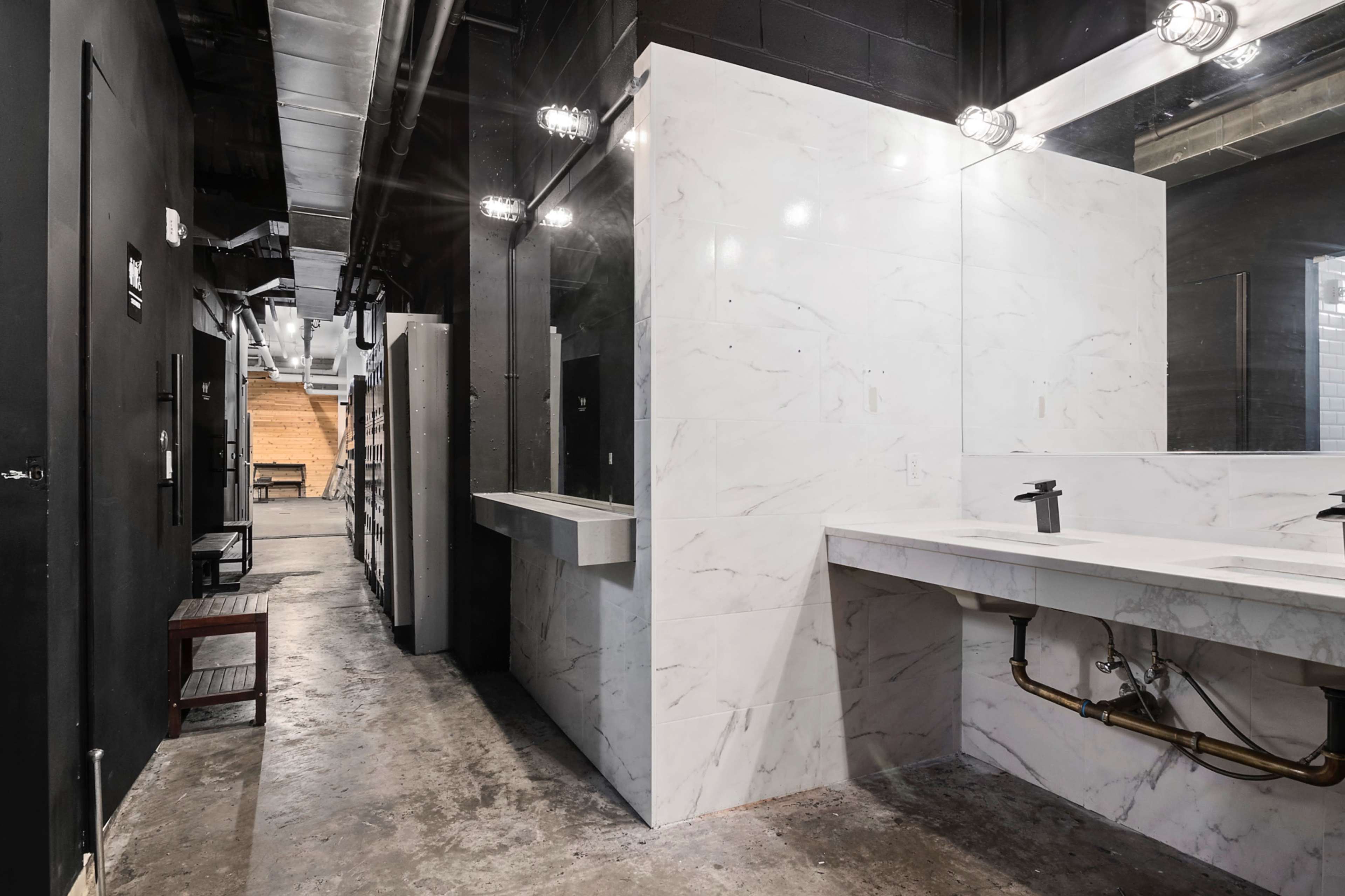 The image shows a modern restroom area with marble walls, a long sink, and industrial lighting along a hallway with a wooden staircase visible in the background.