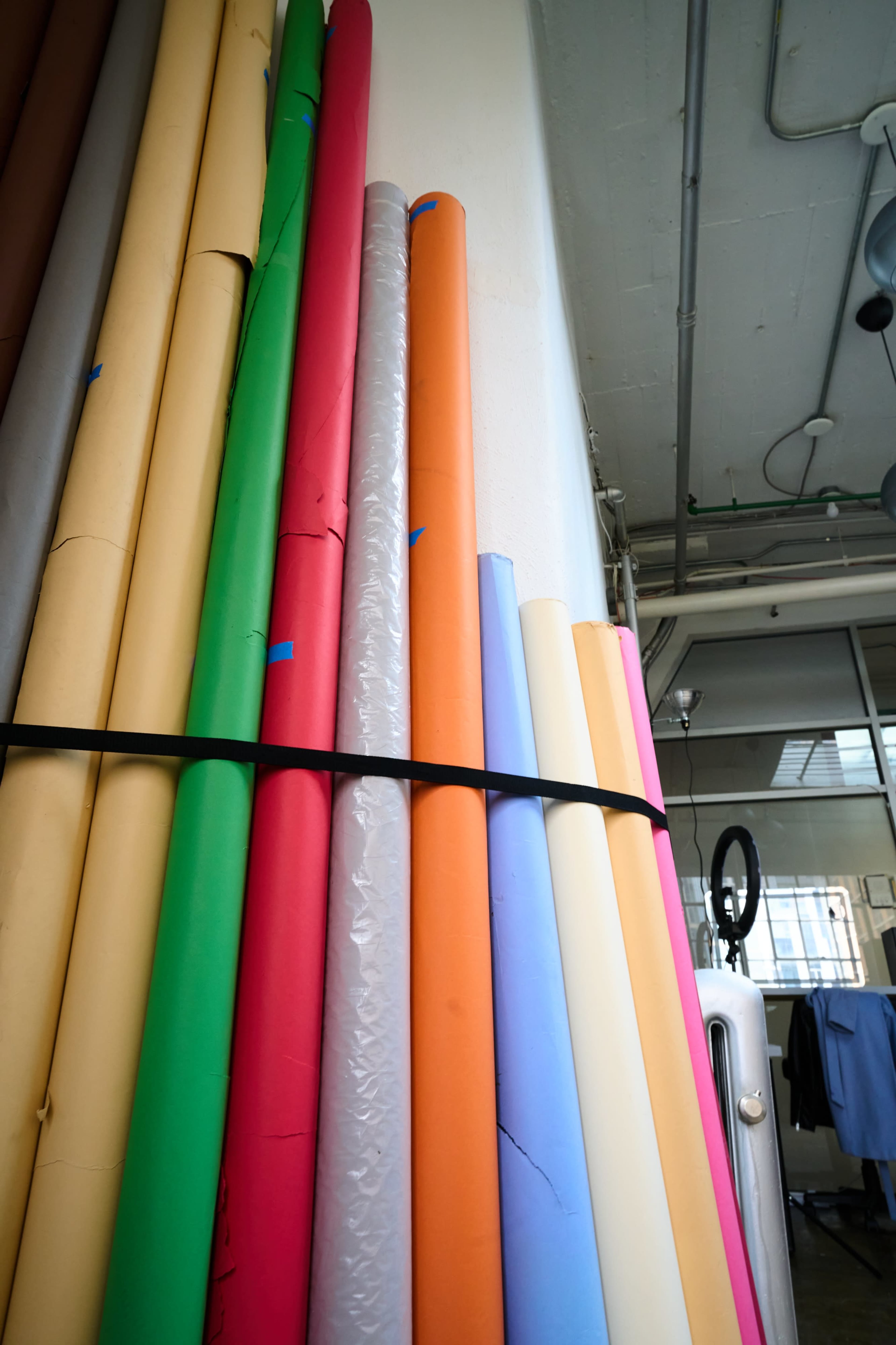 A stack of large rolls of colored photography backdrops is secured with a black strap against a wall.
