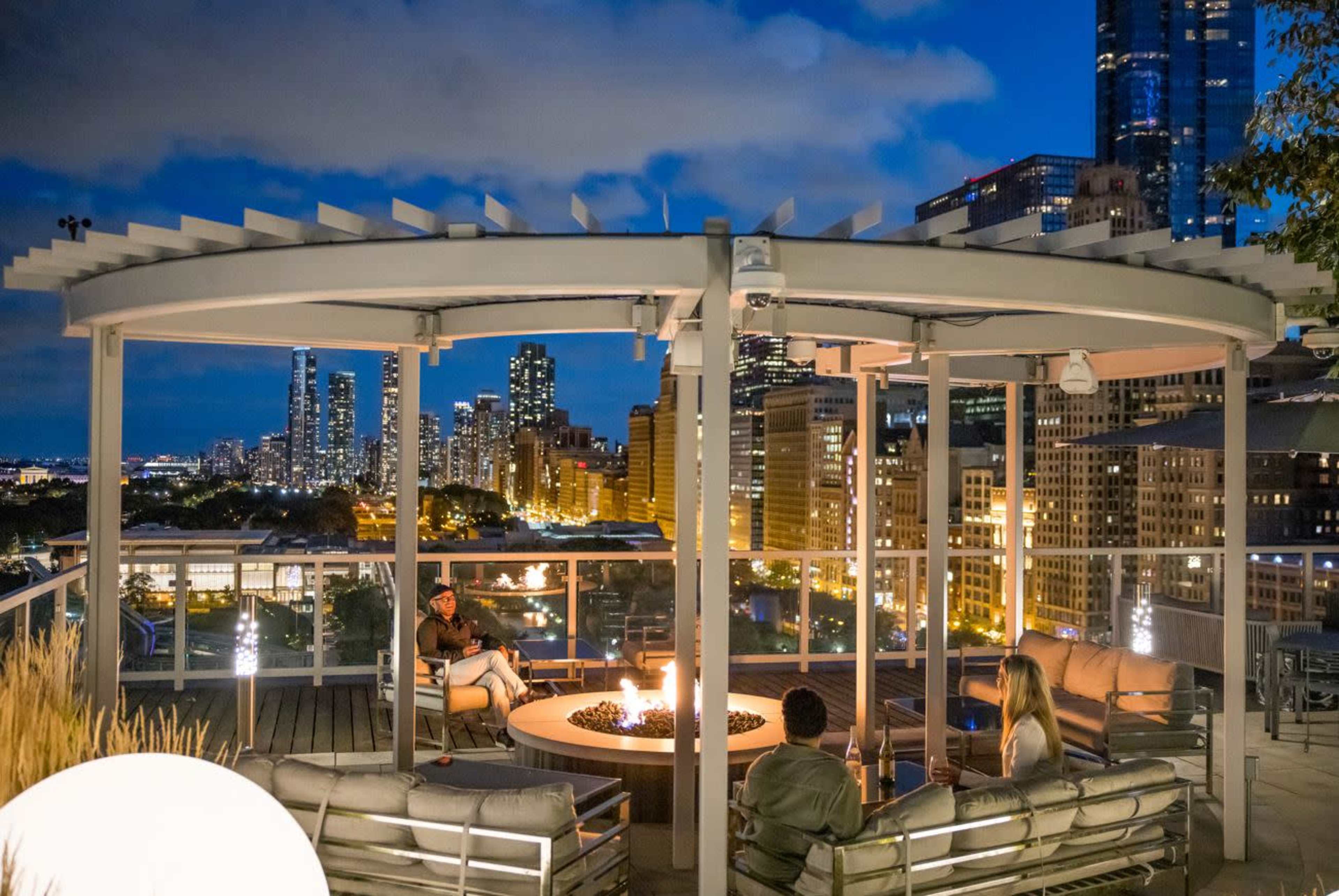 A group of people sit around a fire pit under a pergola on a rooftop, overlooking a city skyline at night.