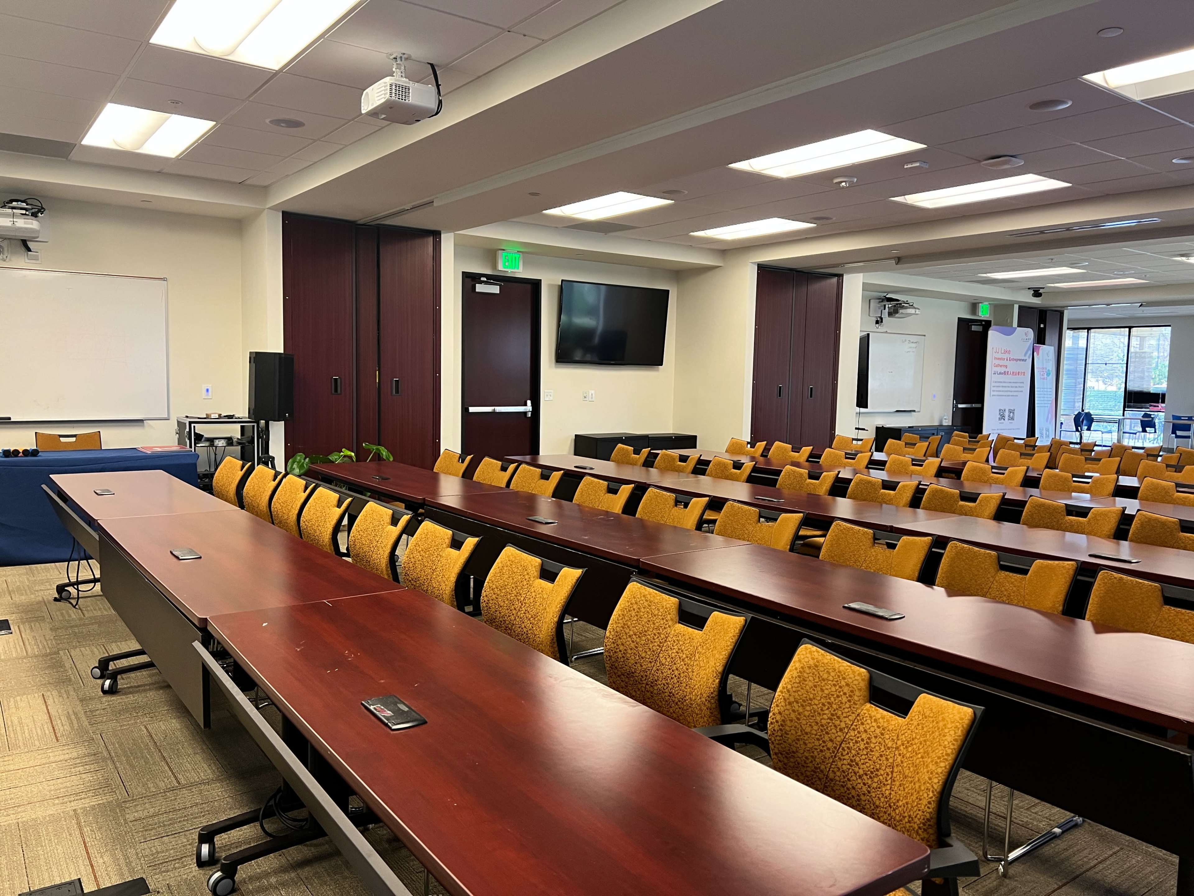 A conference room with multiple rows of wooden tables and yellow chairs, equipped with a projector and screens for presentations.