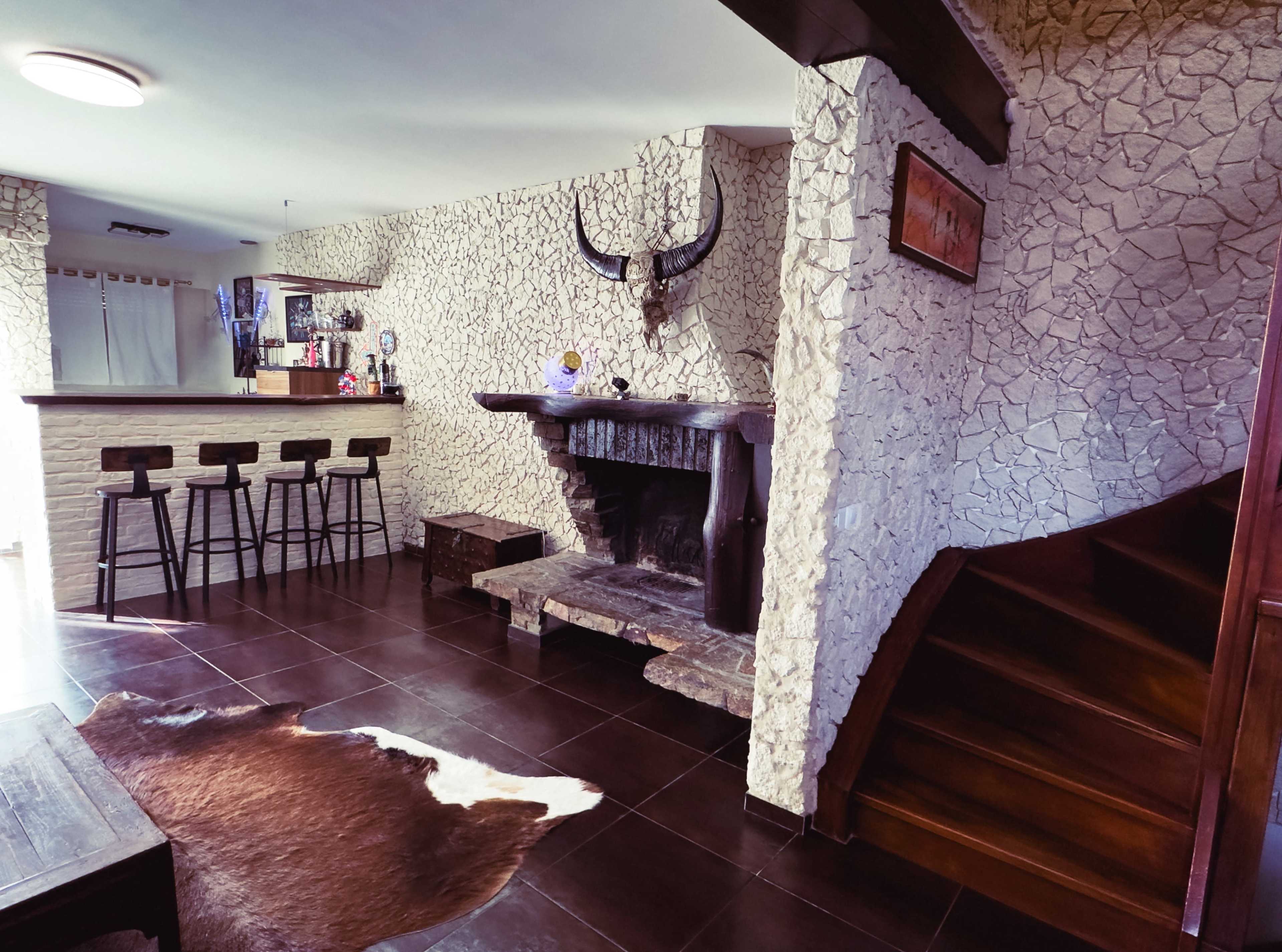 The image shows a living space featuring a stone fireplace, a bar with four stools, and a wooden staircase, all set against textured walls and a dark tile floor.