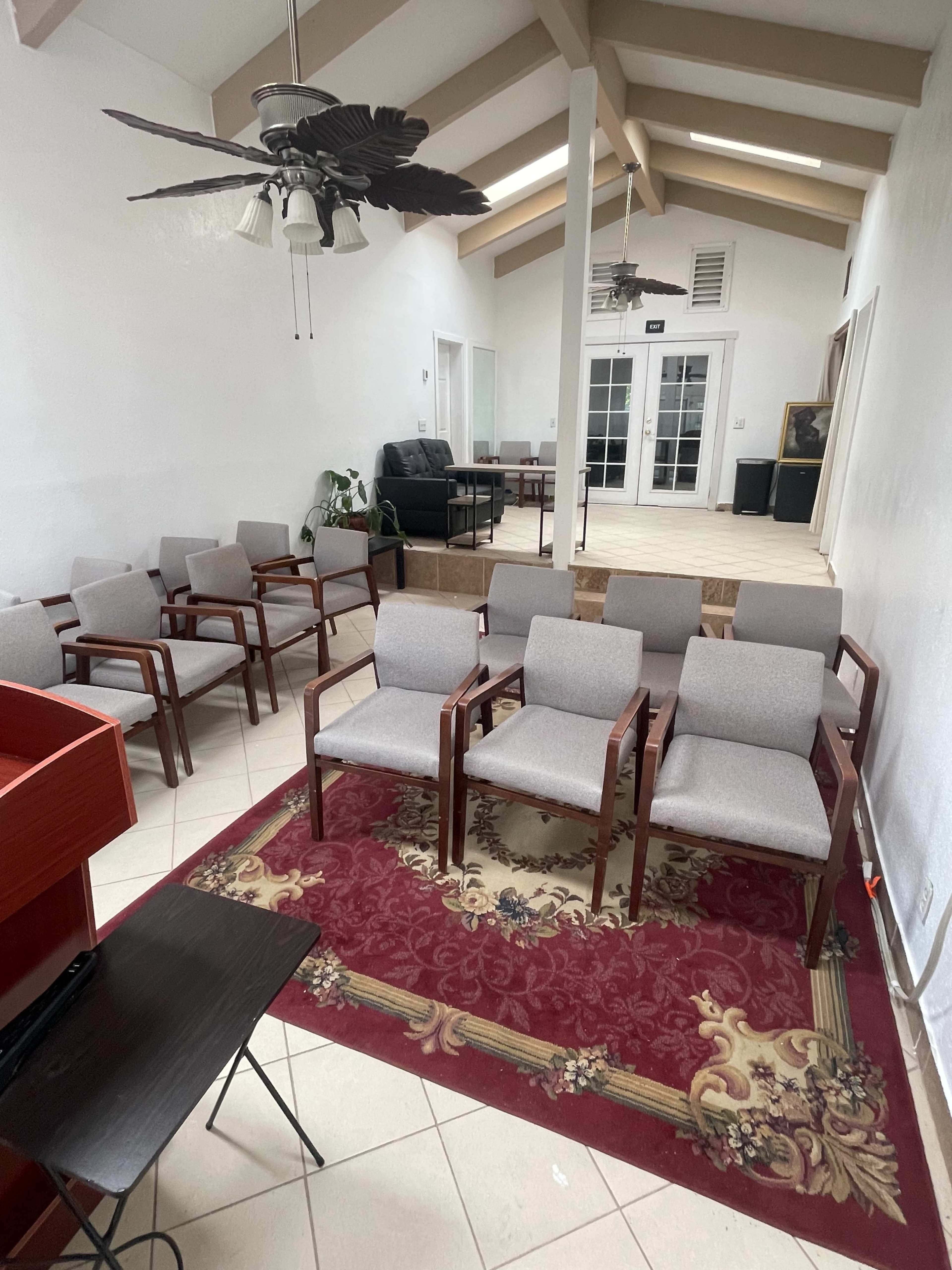 The image shows a room with several rows of gray chairs arranged on a patterned rug, along with a small table and a ceiling fan, all under a vaulted ceiling.
