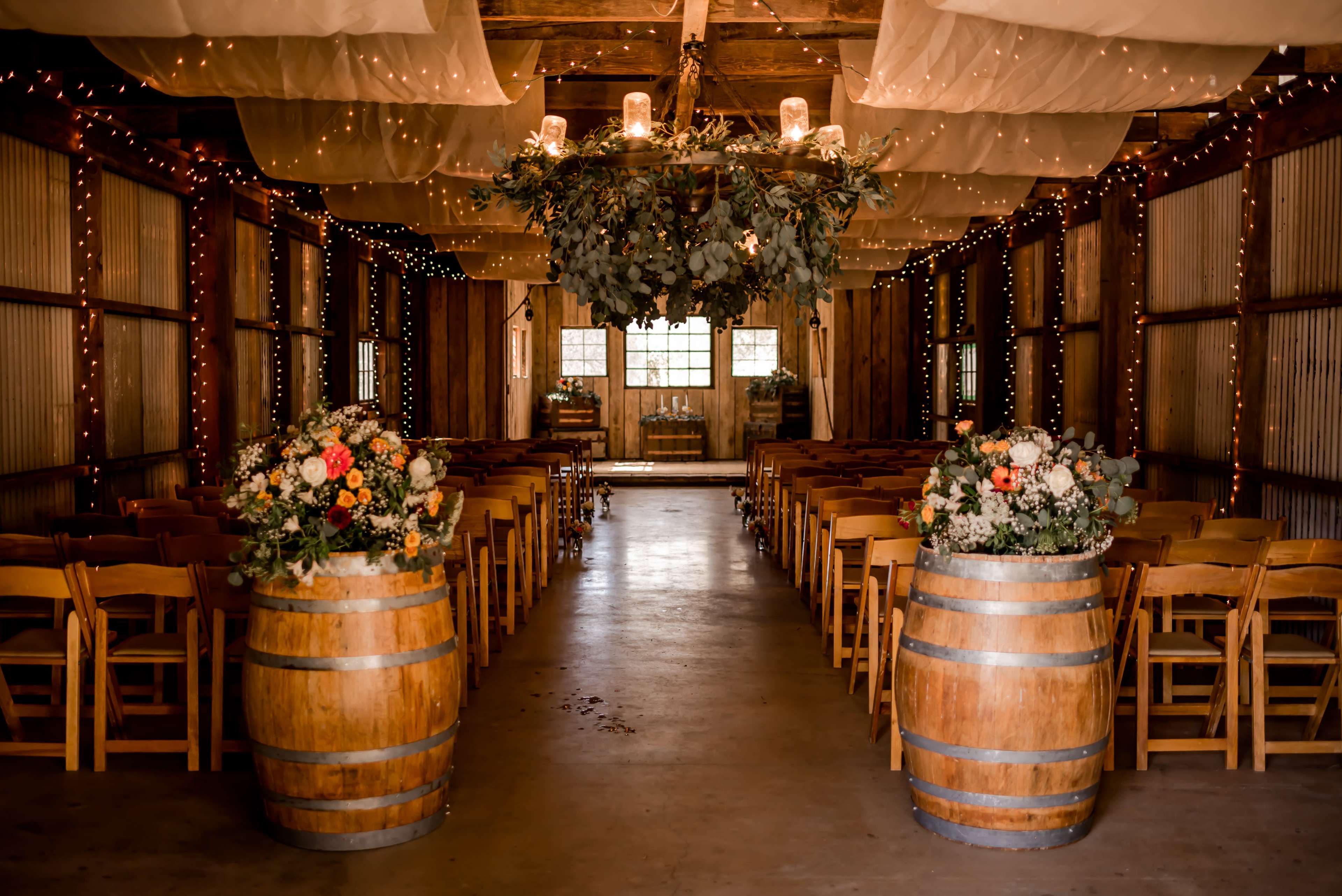 The image shows a rustic indoor venue with wooden barrels filled with flowers at the entrance, leading to rows of chairs and a decorated overhead arrangement.