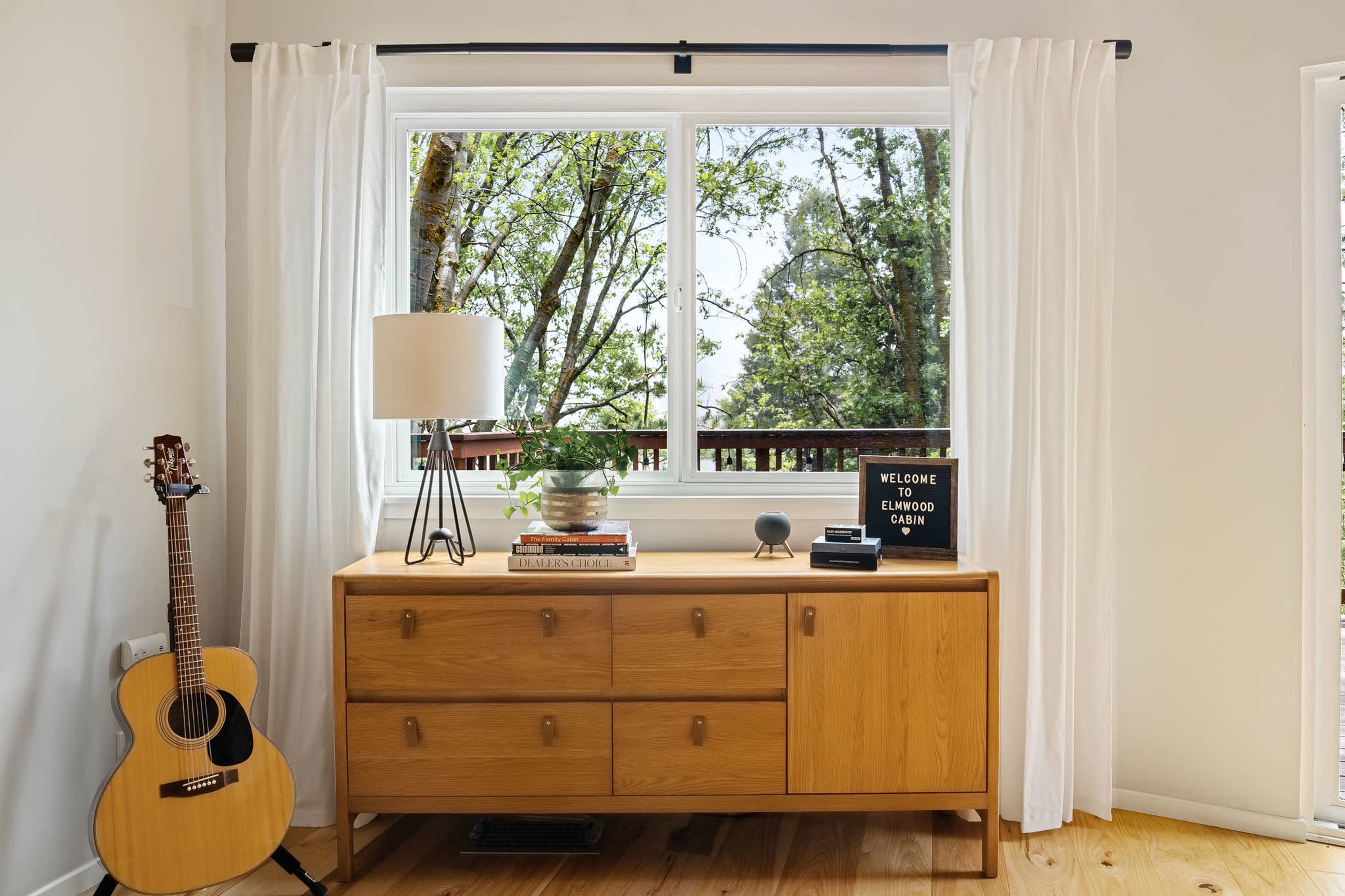 A wooden dresser with a lamp and books stands beside a guitar, facing a window framed by white curtains that overlooks a view of trees.