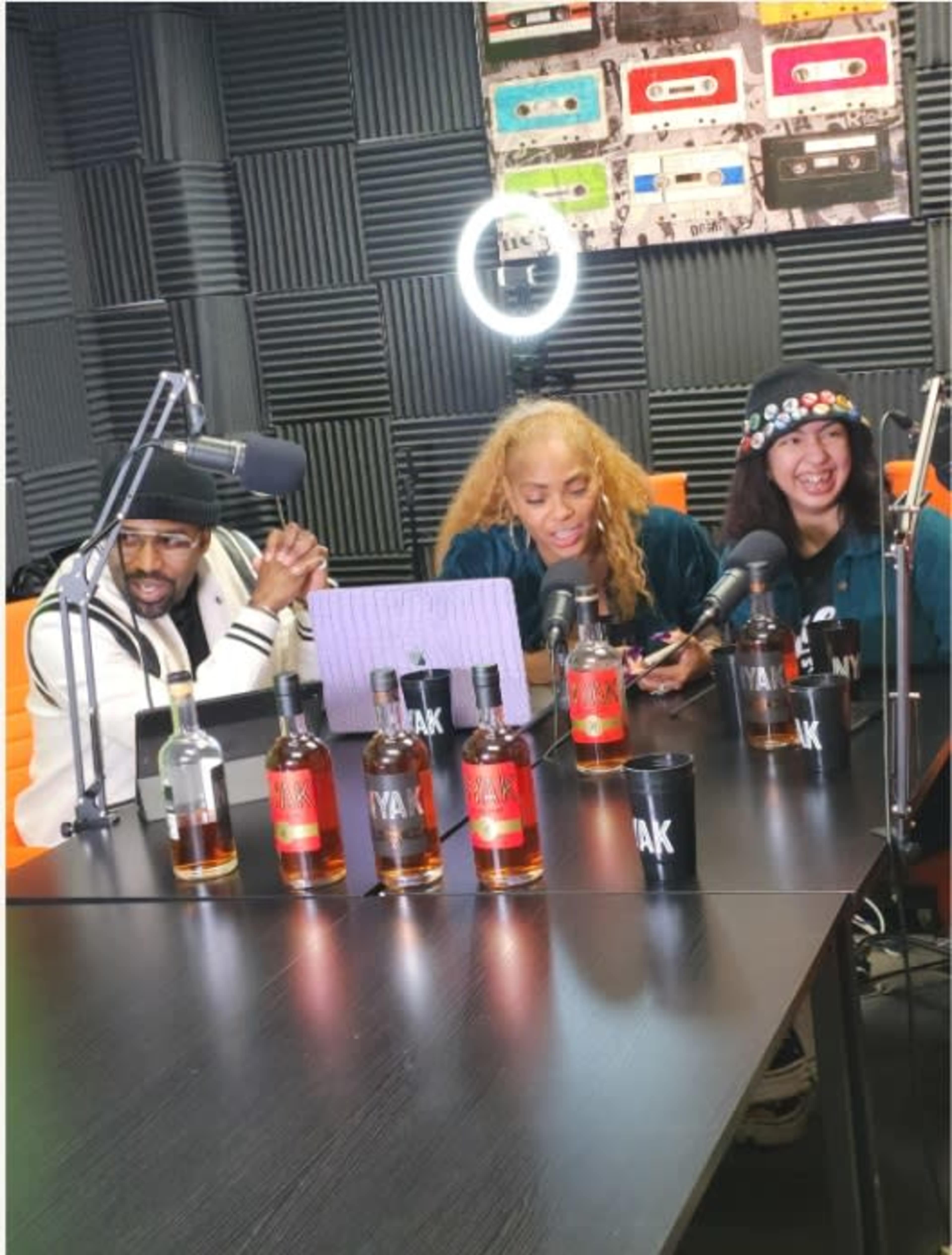 Three individuals are seated at a table in a recording studio, with several bottles of liquor and a laptop in front of them.
