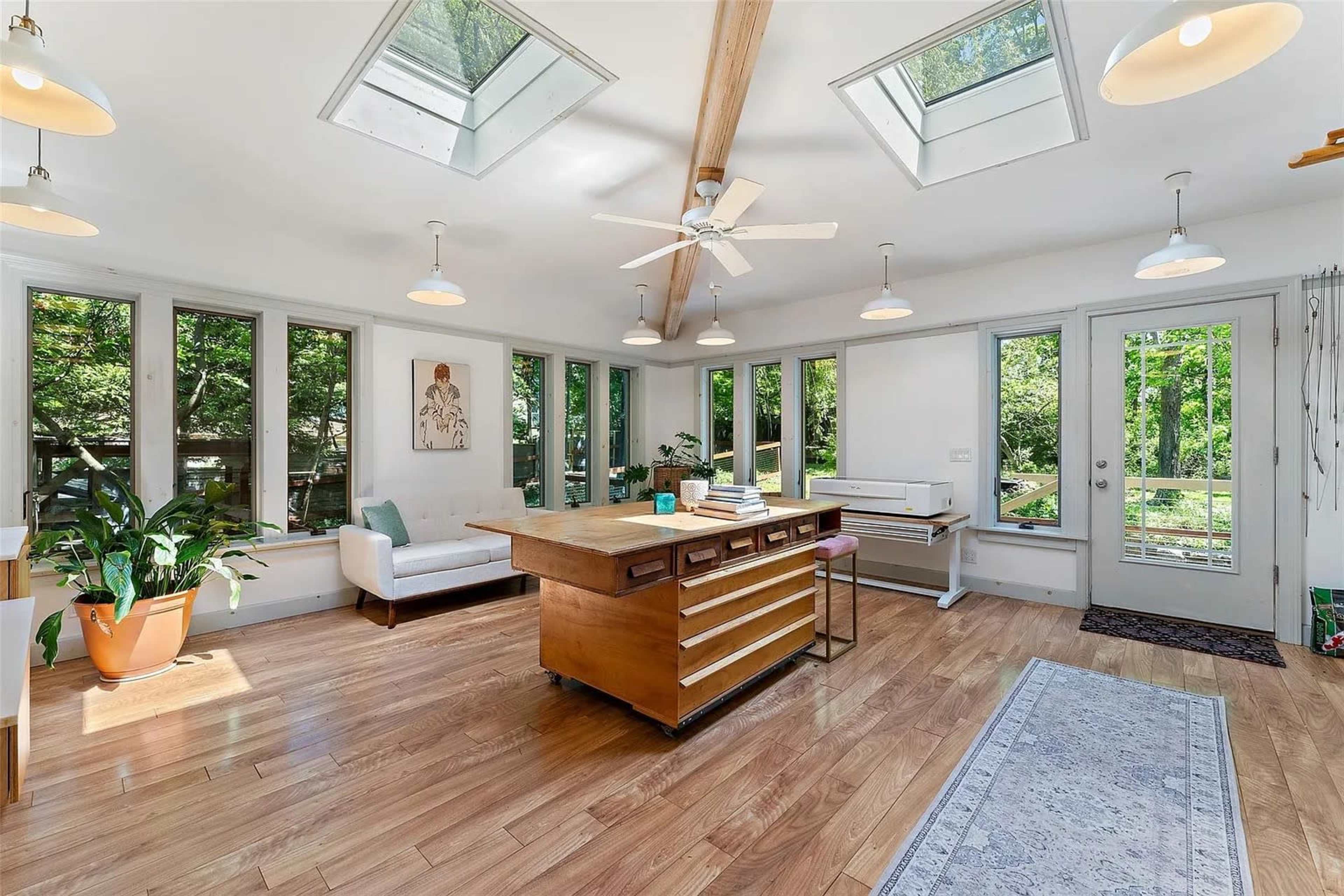 The image shows a bright, spacious room featuring large windows, a central wooden island with drawers, a ceiling fan, and plants, leading to an outdoor view.