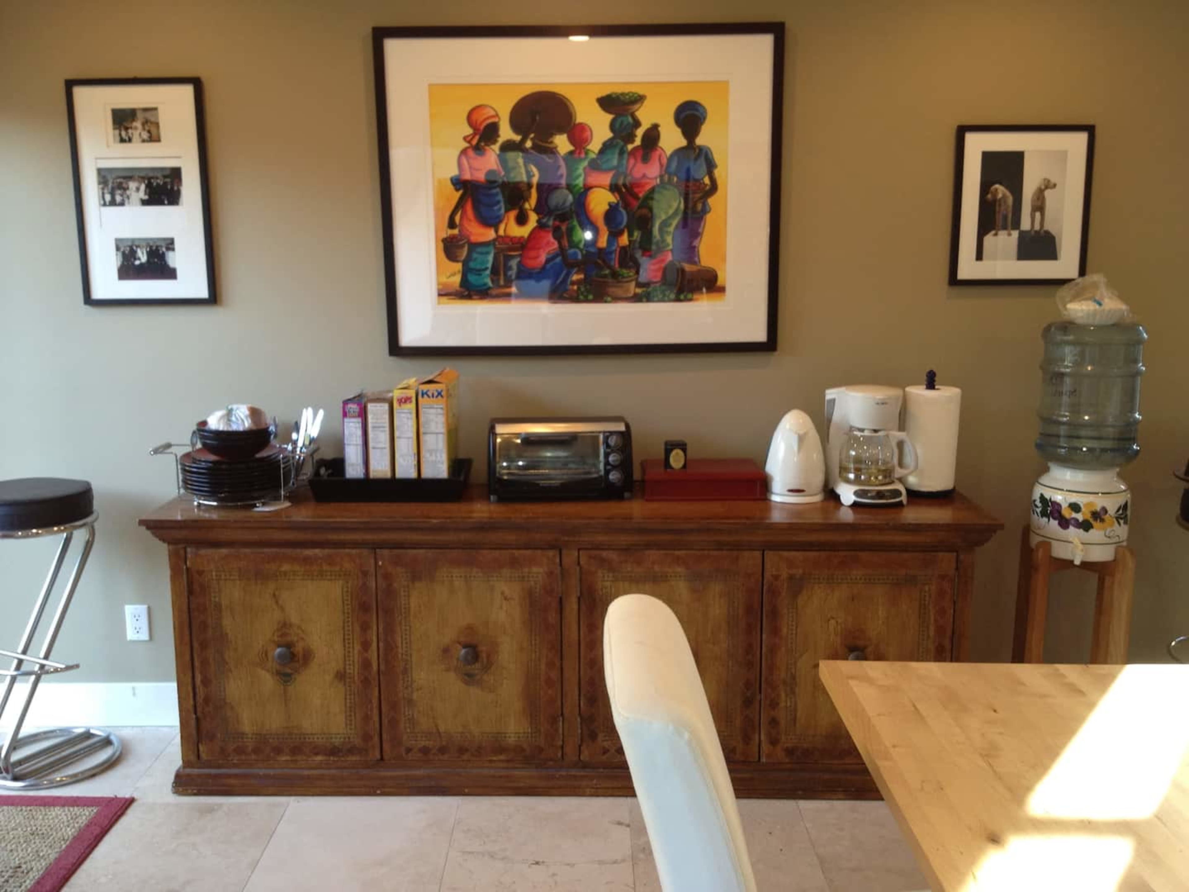 A wooden sideboard adorned with various kitchen appliances and an art piece featuring colorful figures, set against a wall with framed photographs.