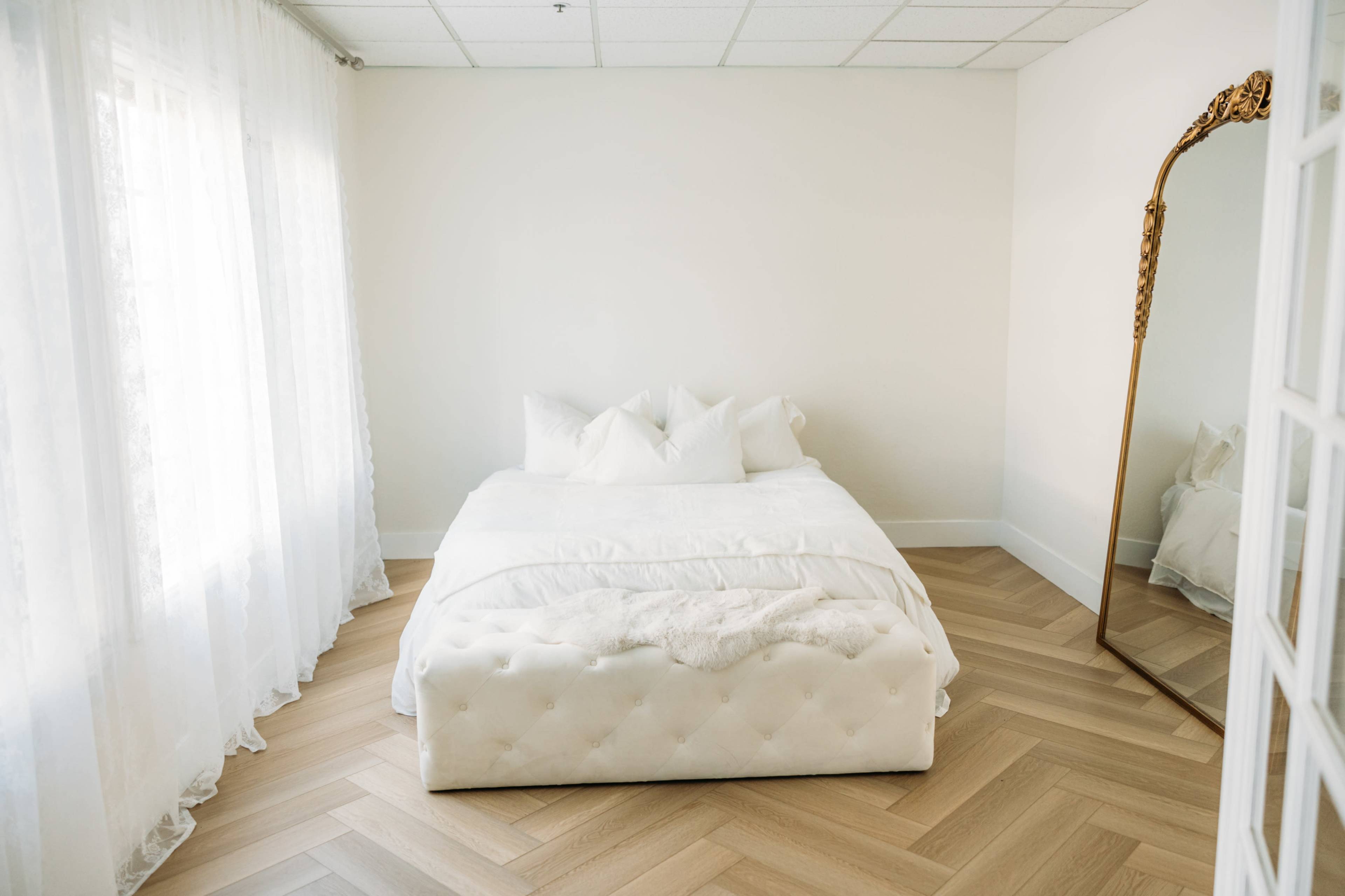 A minimalistic bedroom features a white bed with pillows, a large mirror, and hardwood flooring, illuminated by natural light from sheer curtains.