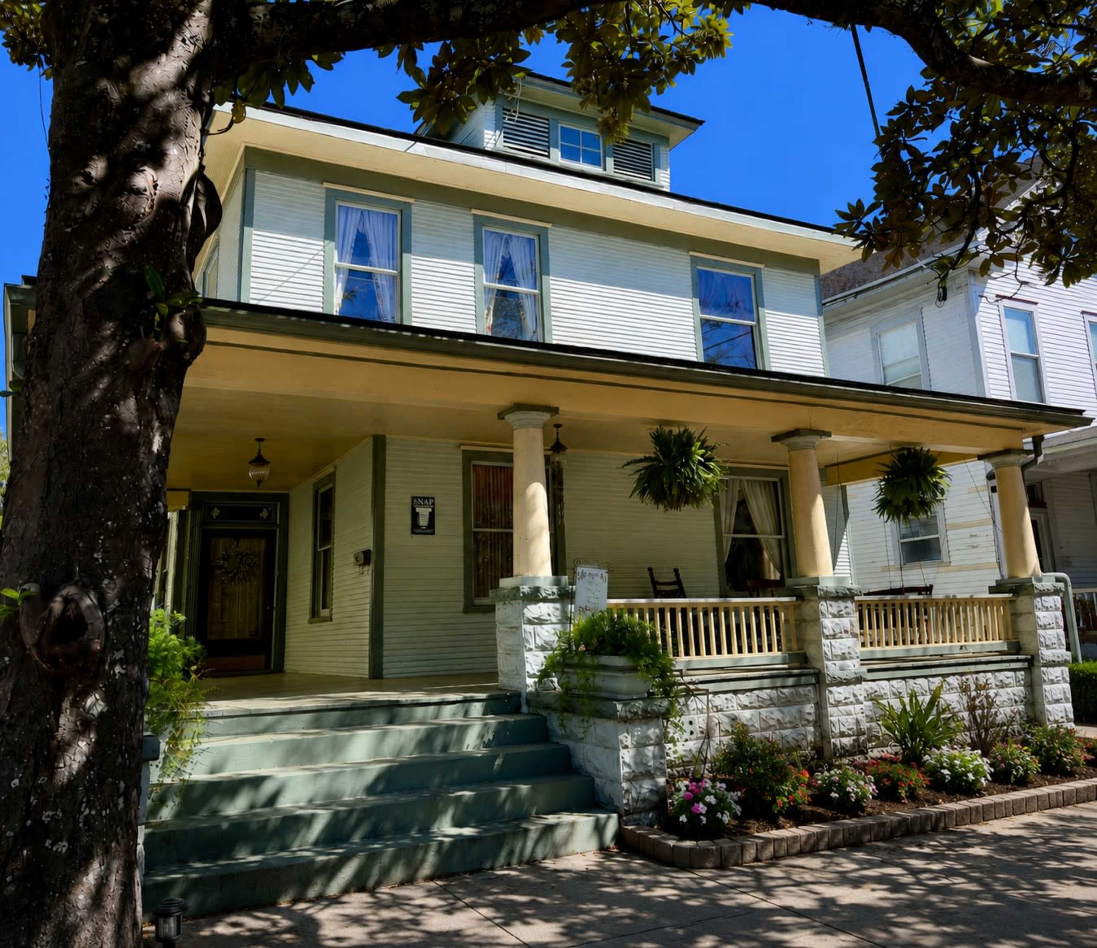 The image shows a two-story house with a wraparound porch, supported by stone columns, and surrounded by landscaped gardens and trees.