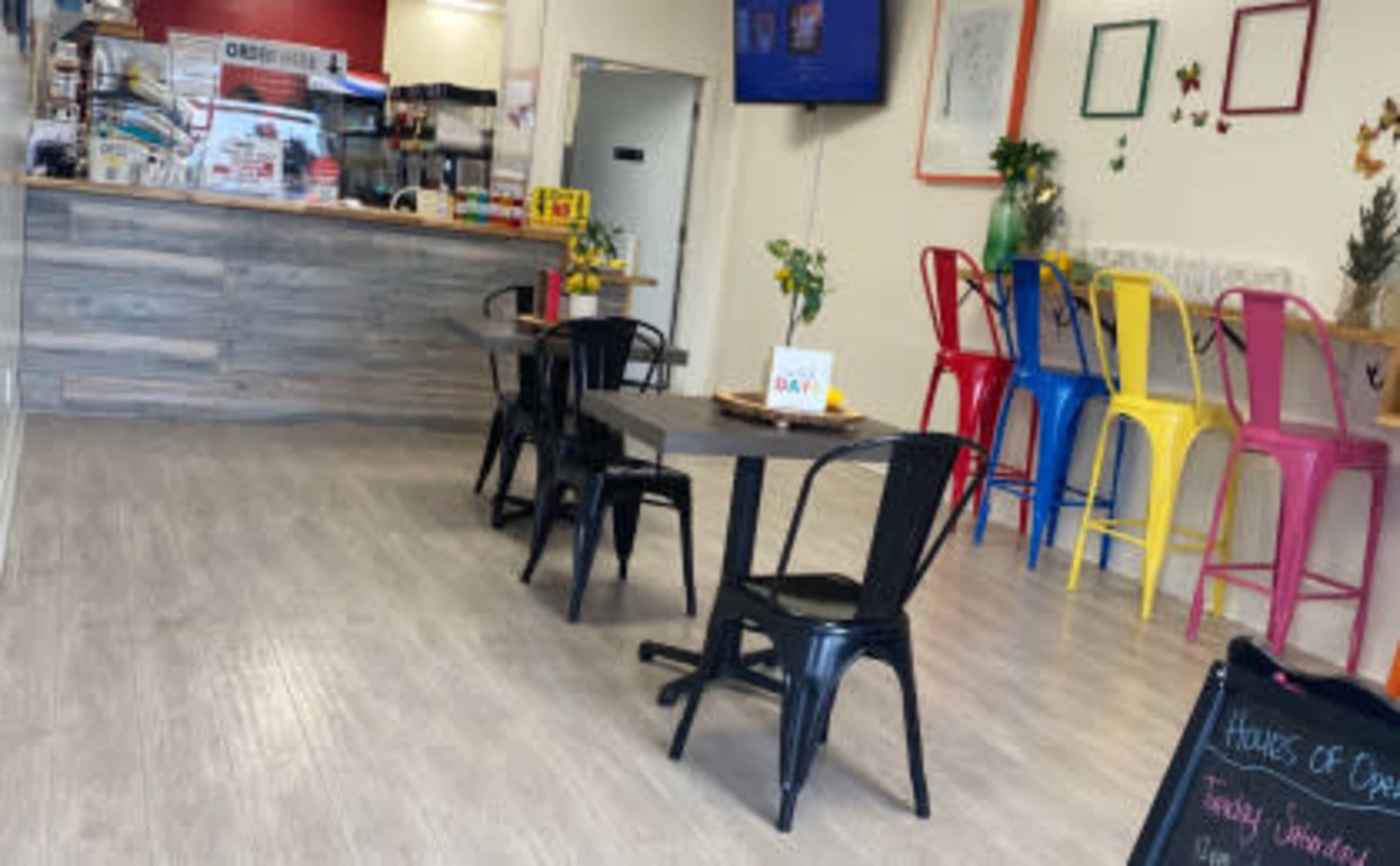 The image shows a bright, modern café with a service counter, a television on the wall, and colorful metal chairs around several black tables.