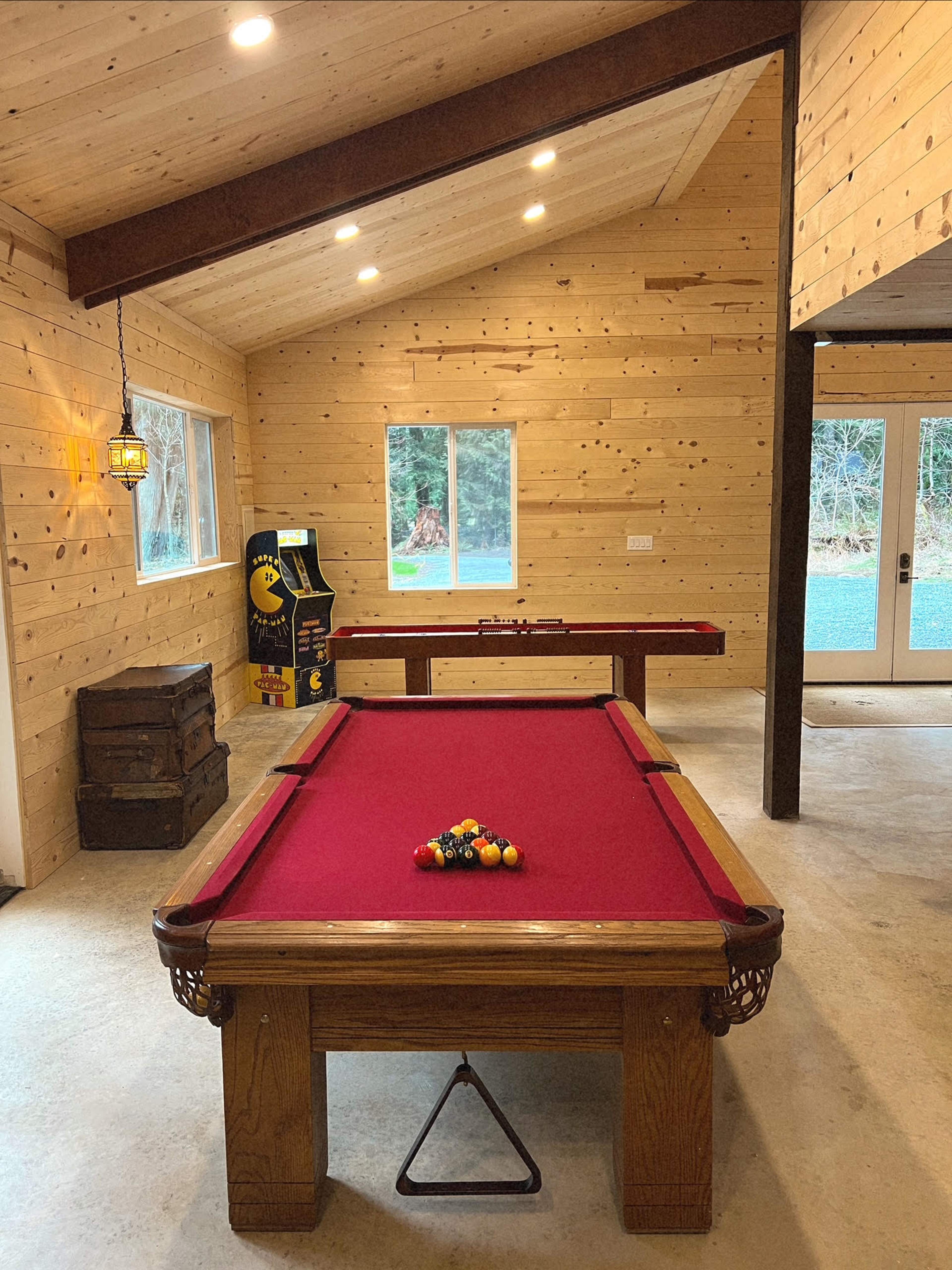 A game room featuring a red felt pool table, a wooden air hockey table, and a vintage Pac-Man arcade machine, all set against a backdrop of rustic wooden walls.