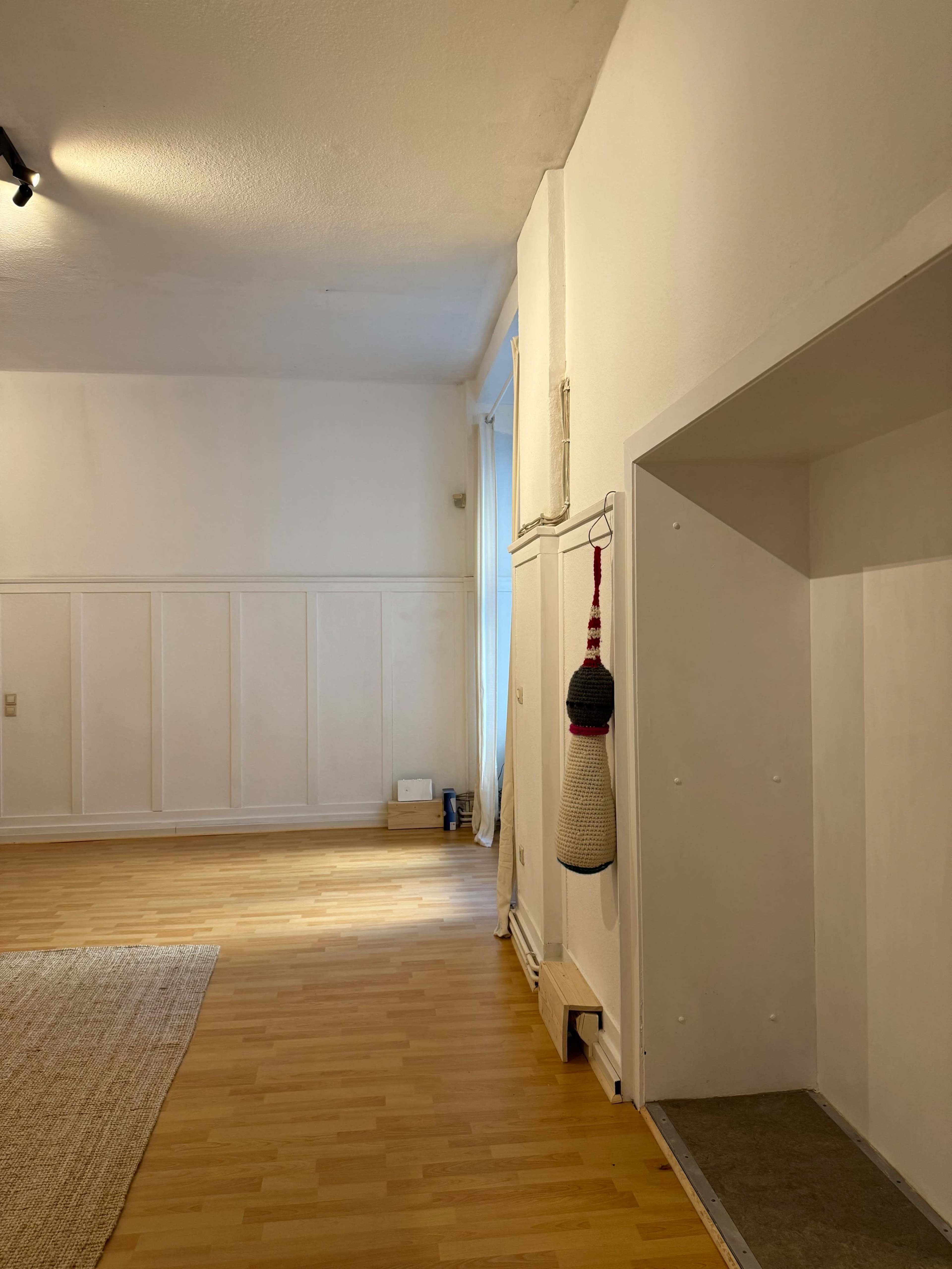 The image shows a spacious, minimalist interior with wooden floors, white walls, and a small alcove on the right side.