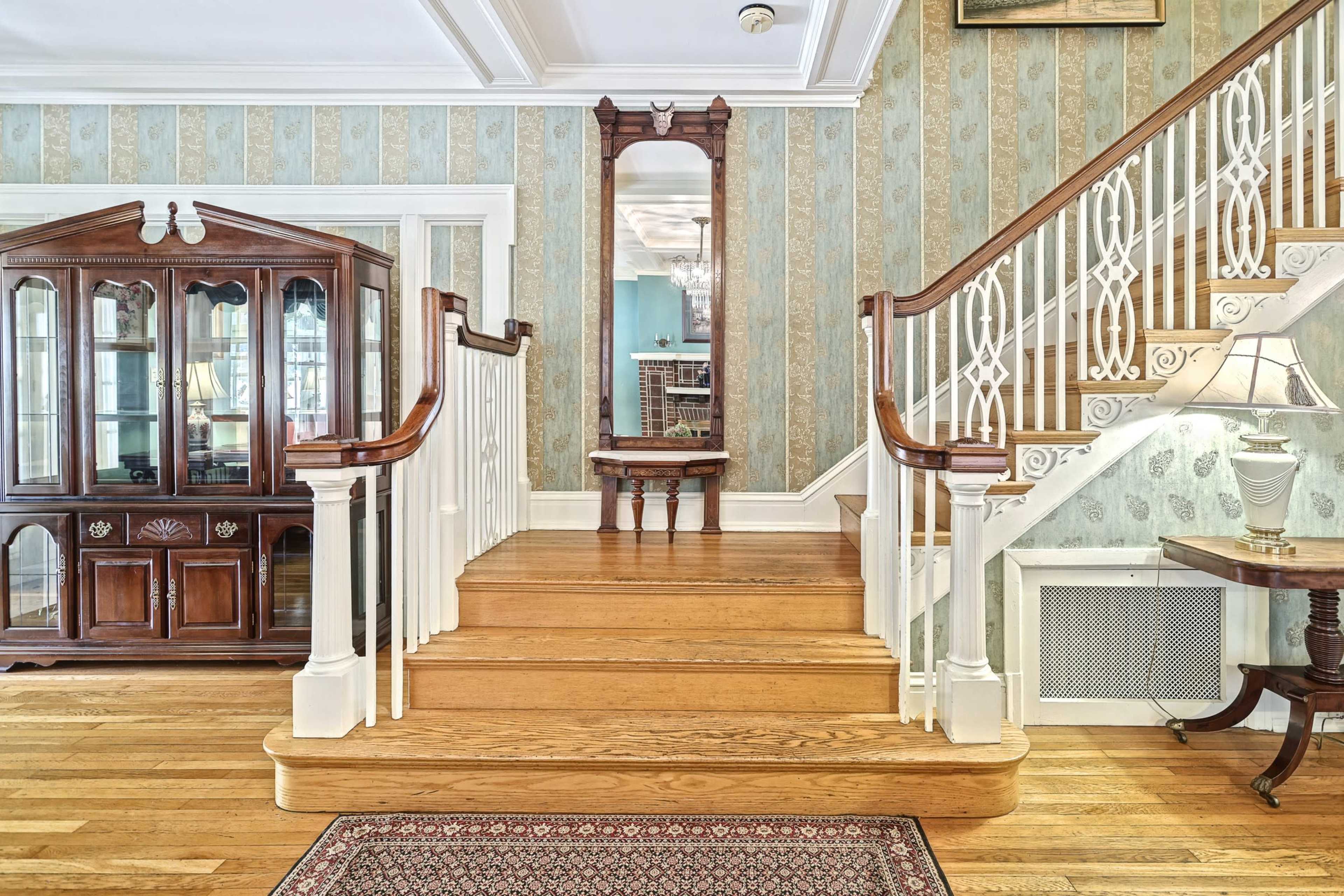The scene features a wooden staircase leading to an upper level, flanked by a glass-fronted cabinet and a decorative mirror on the wall.
