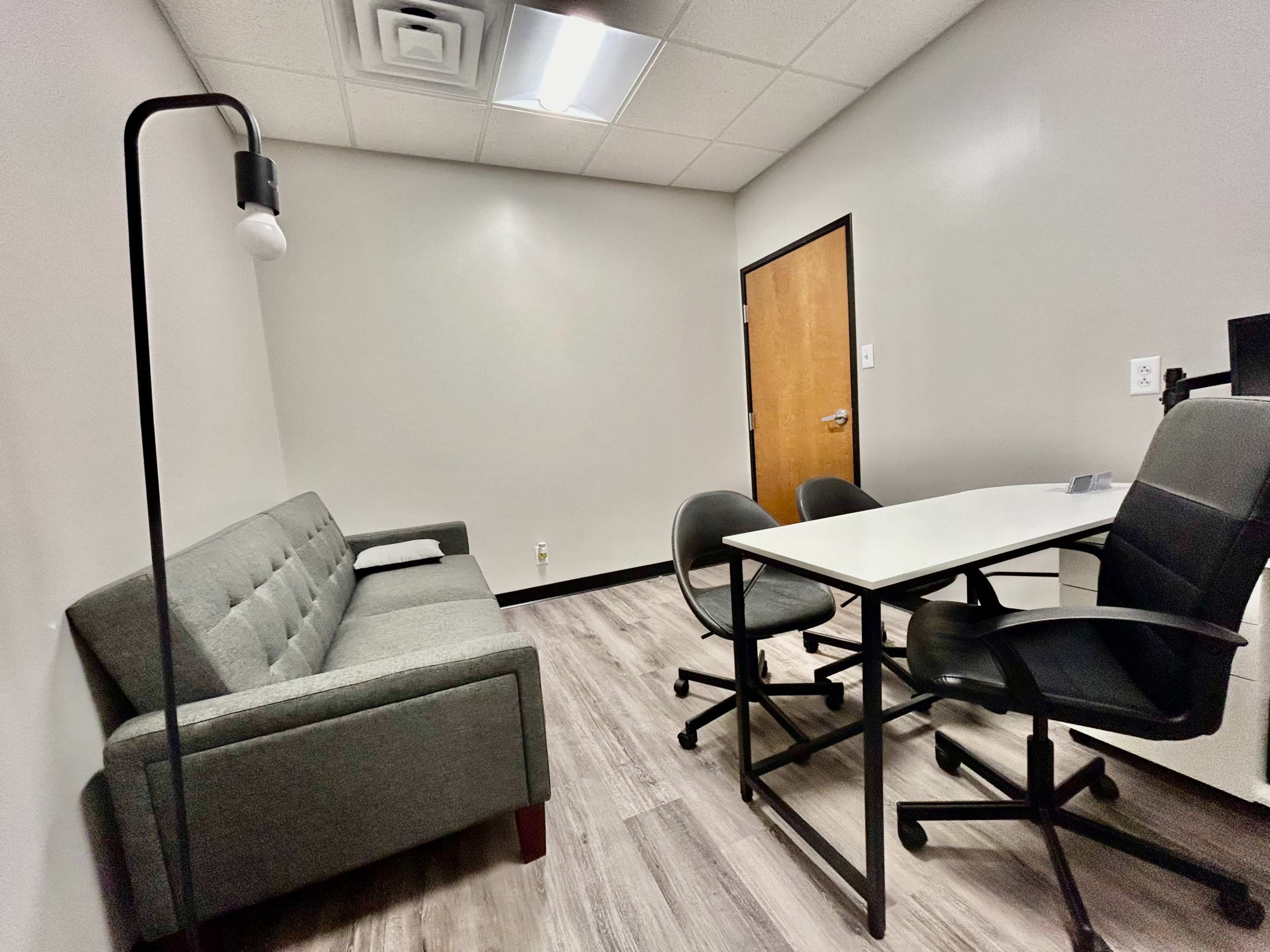 A small, furnished office with a gray couch, two black chairs, and a white table, featuring plain walls and a wooden door.