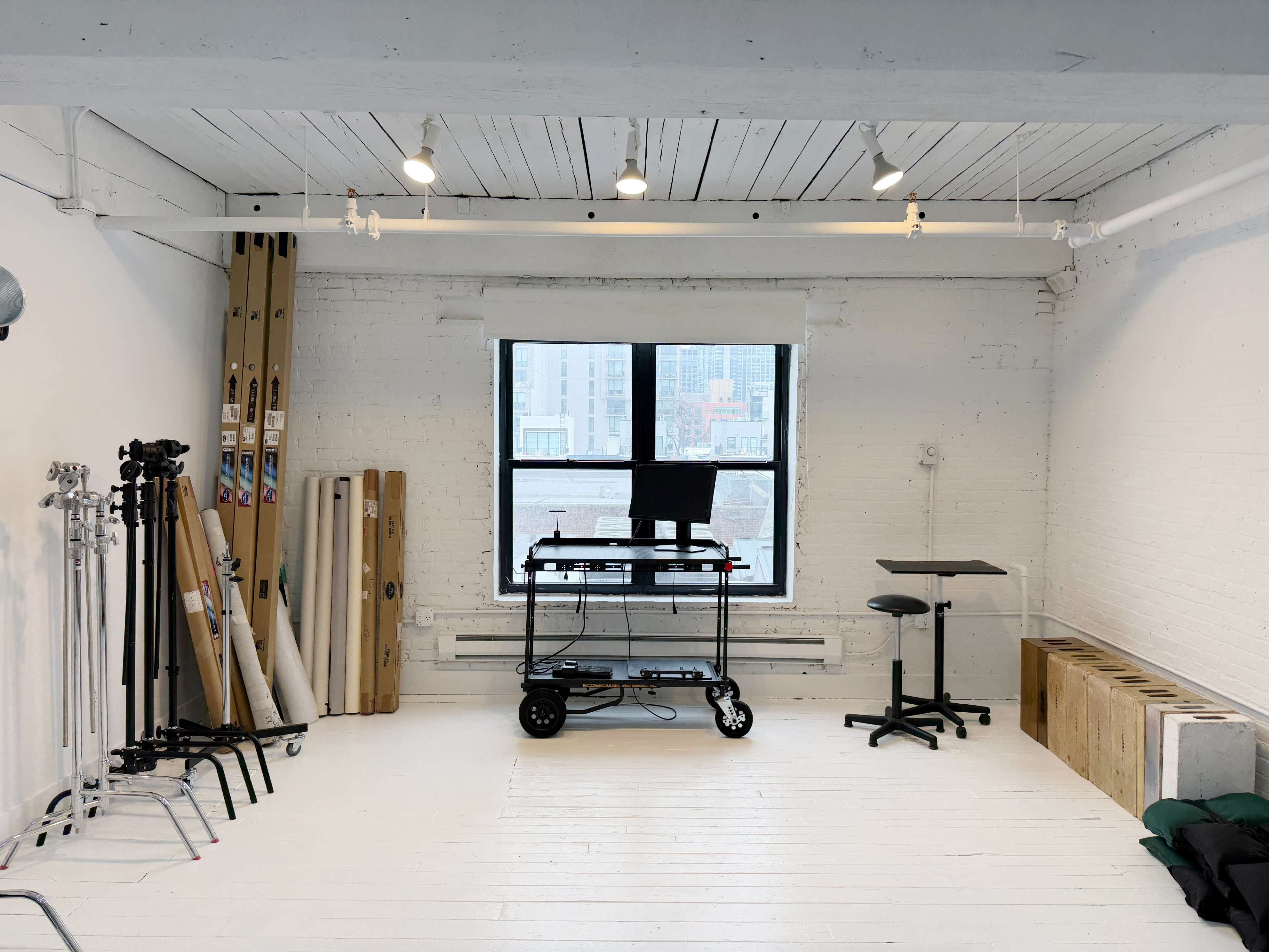 The image shows a spacious, minimally furnished studio with a large window, a desk with a computer, and various equipment arranged along the walls.
