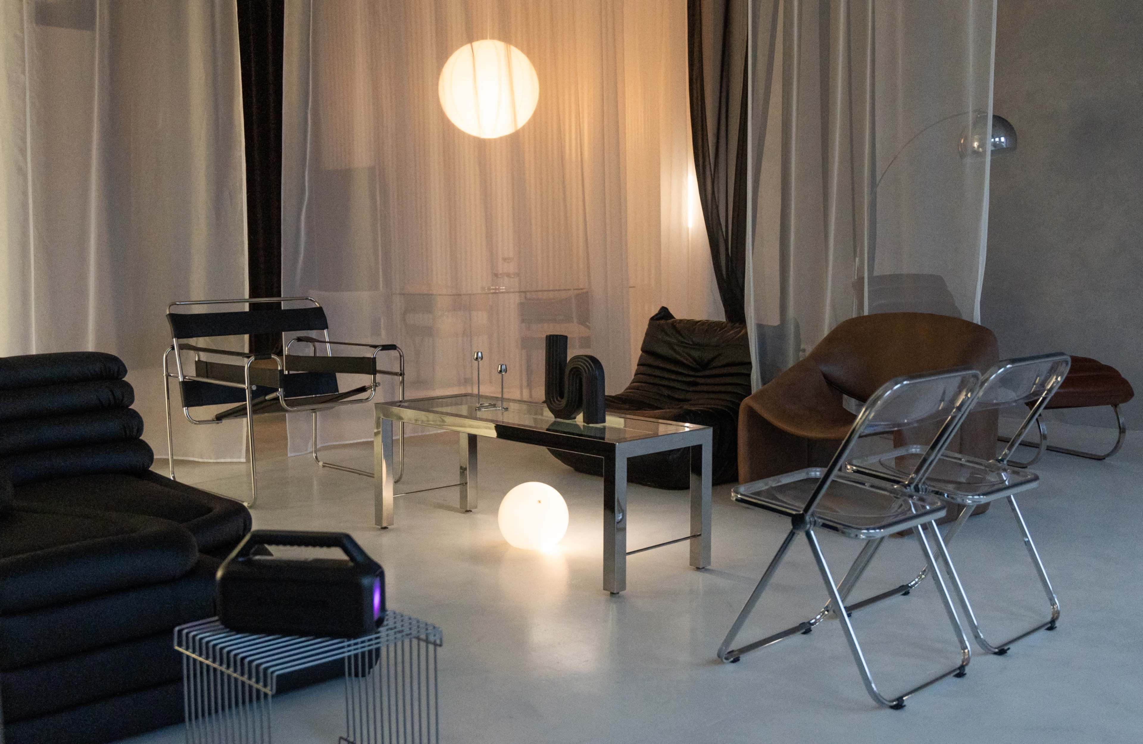 The image shows a modern living room featuring minimalist furniture, including a sleek glass coffee table, black seating, and a prominent light fixture.