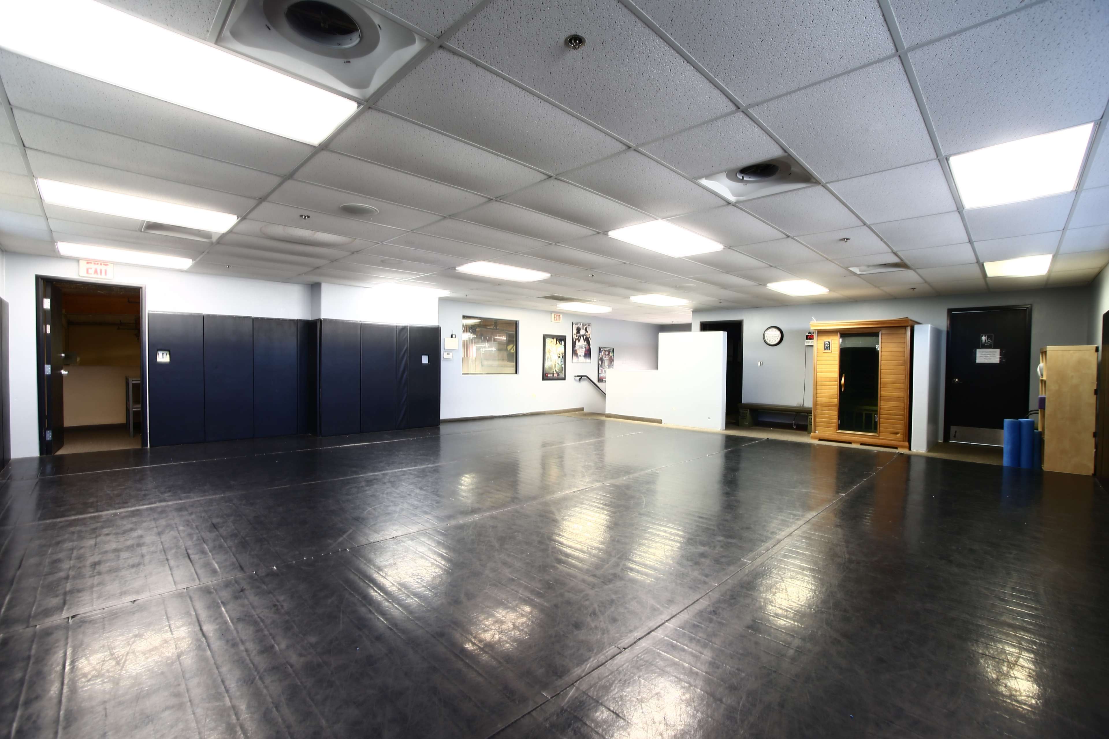 A spacious, empty room with a black rubber flooring, light gray walls, and various wall-mounted features, including a clock and framed pictures.