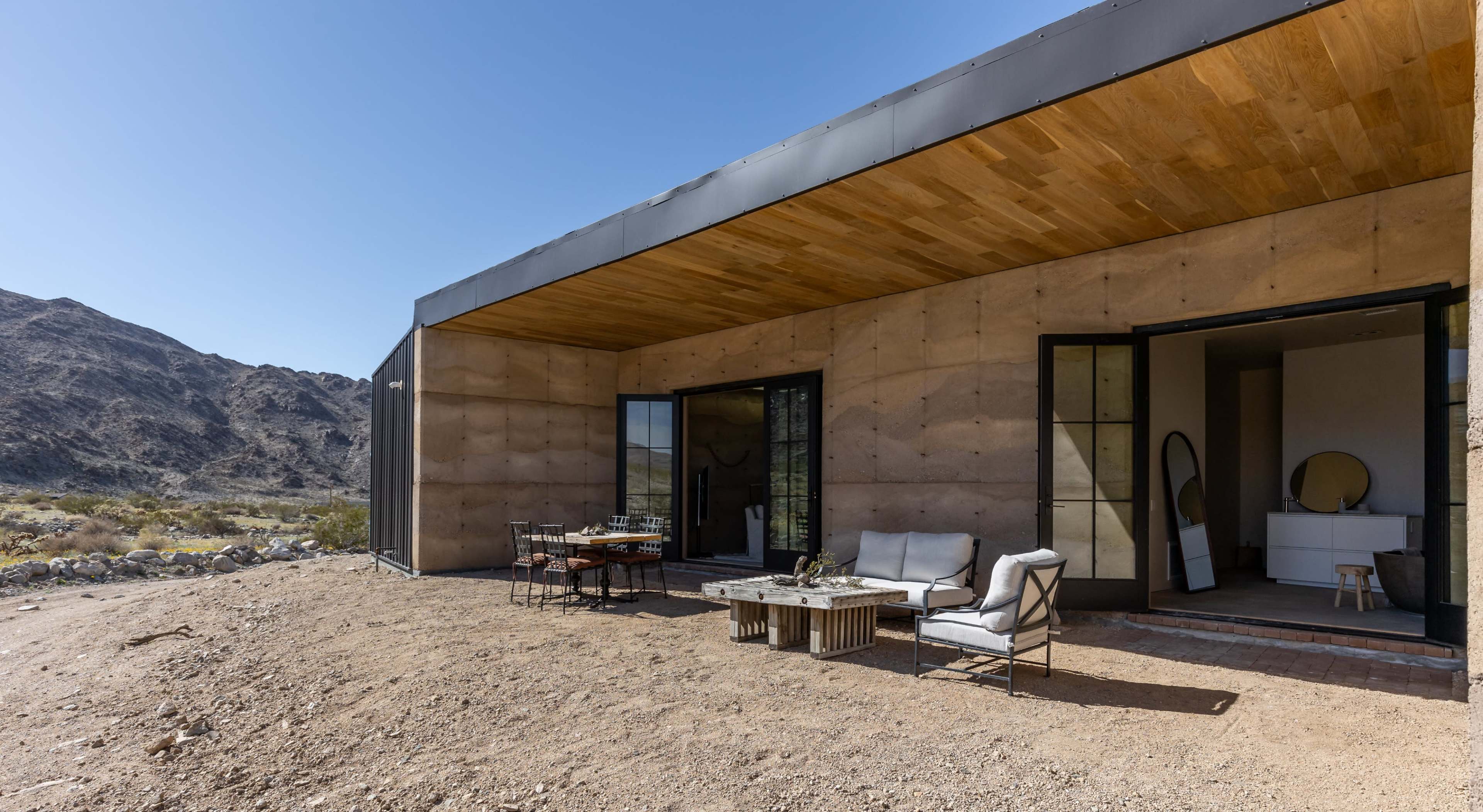 A modern building with a concrete facade and large glass doors overlooks a desert landscape, featuring outdoor seating and a dining area.