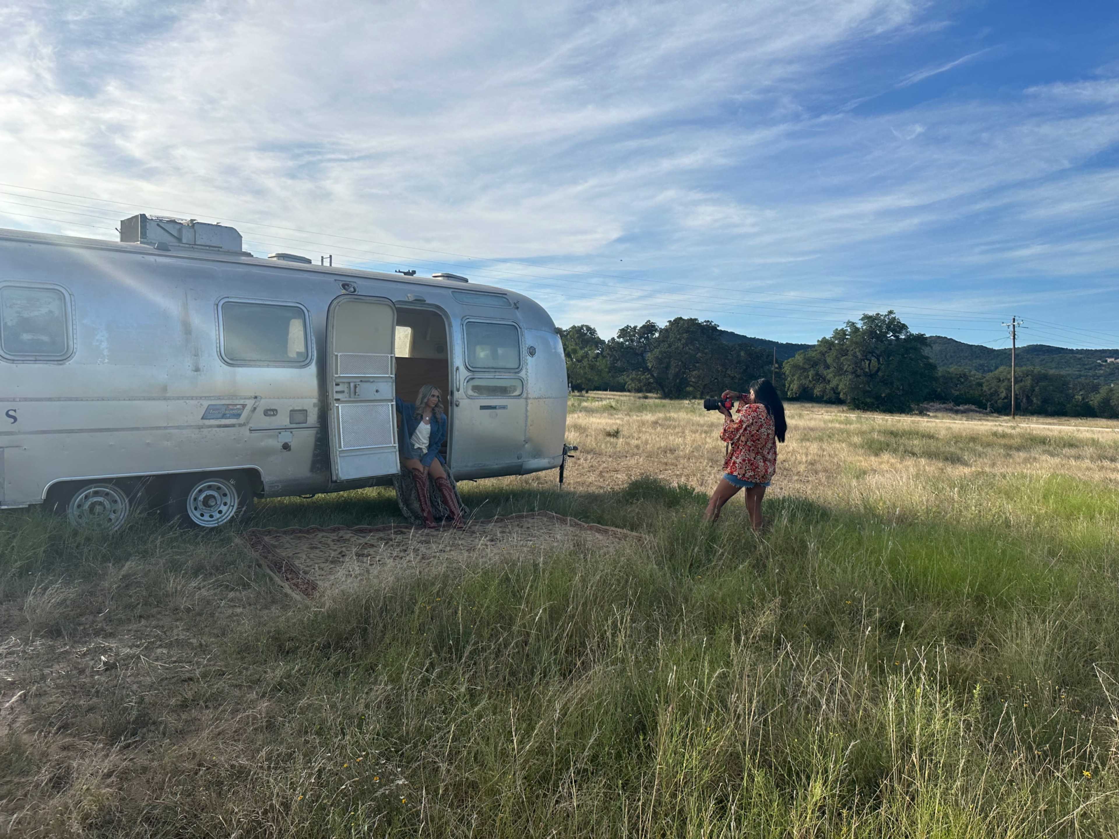Two individuals are engaged in a photo shoot near a vintage Airstream trailer in a grassy field under a blue sky.