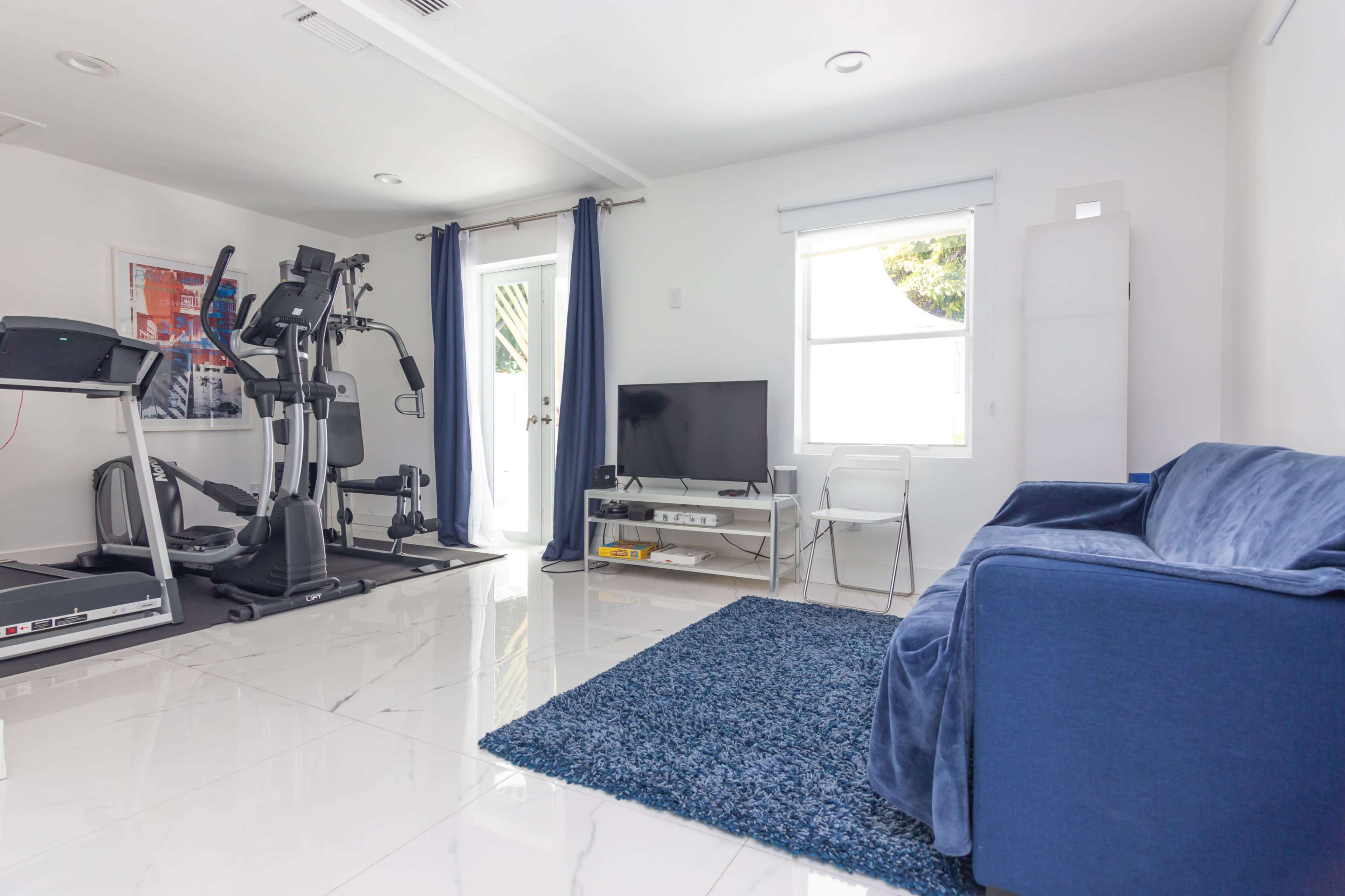 The room features a treadmill and exercise equipment beside a blue couch, a TV on a white stand, and a door leading outside.