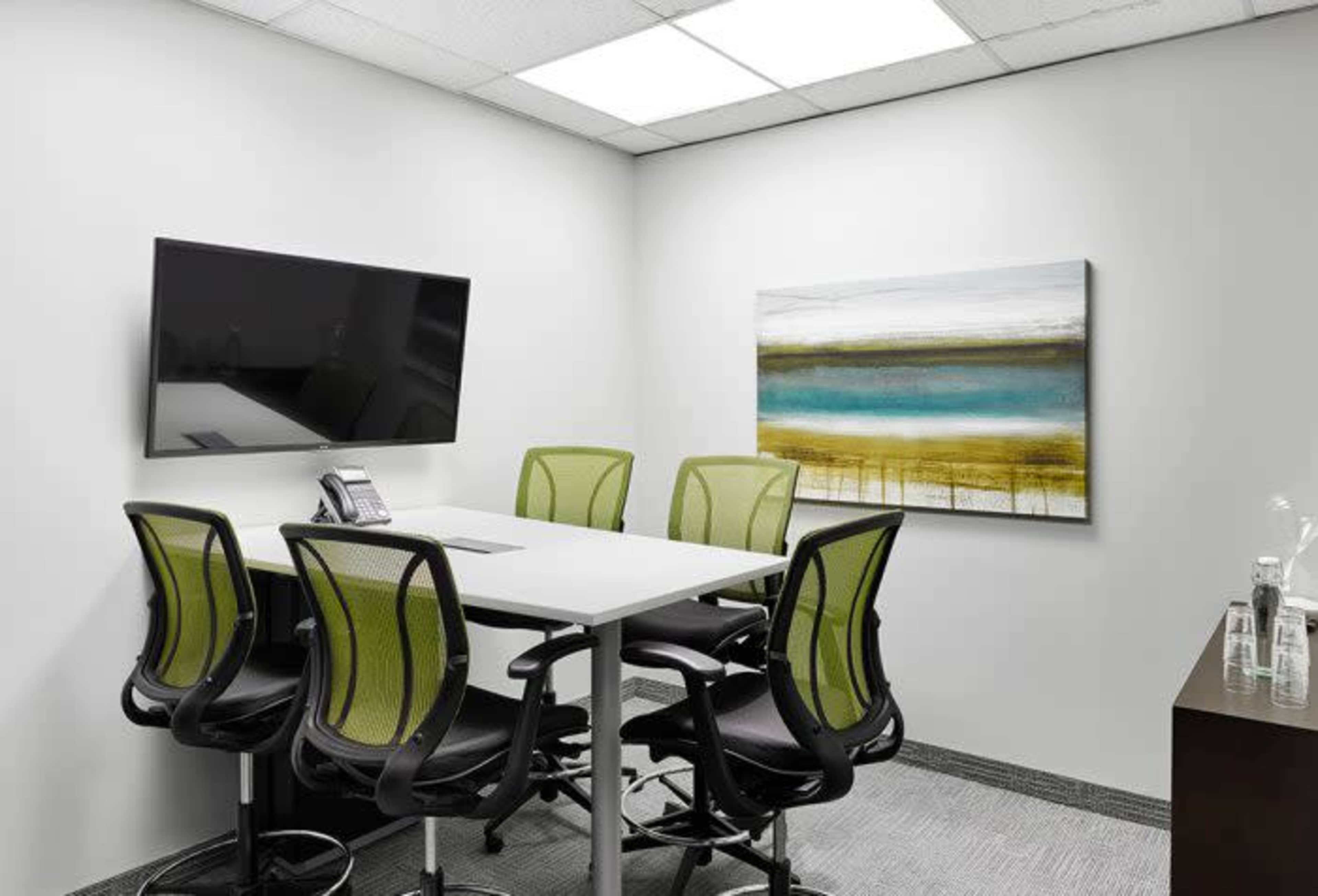 A small conference room with a table surrounded by five green office chairs, a wall-mounted TV, and an abstract painting.