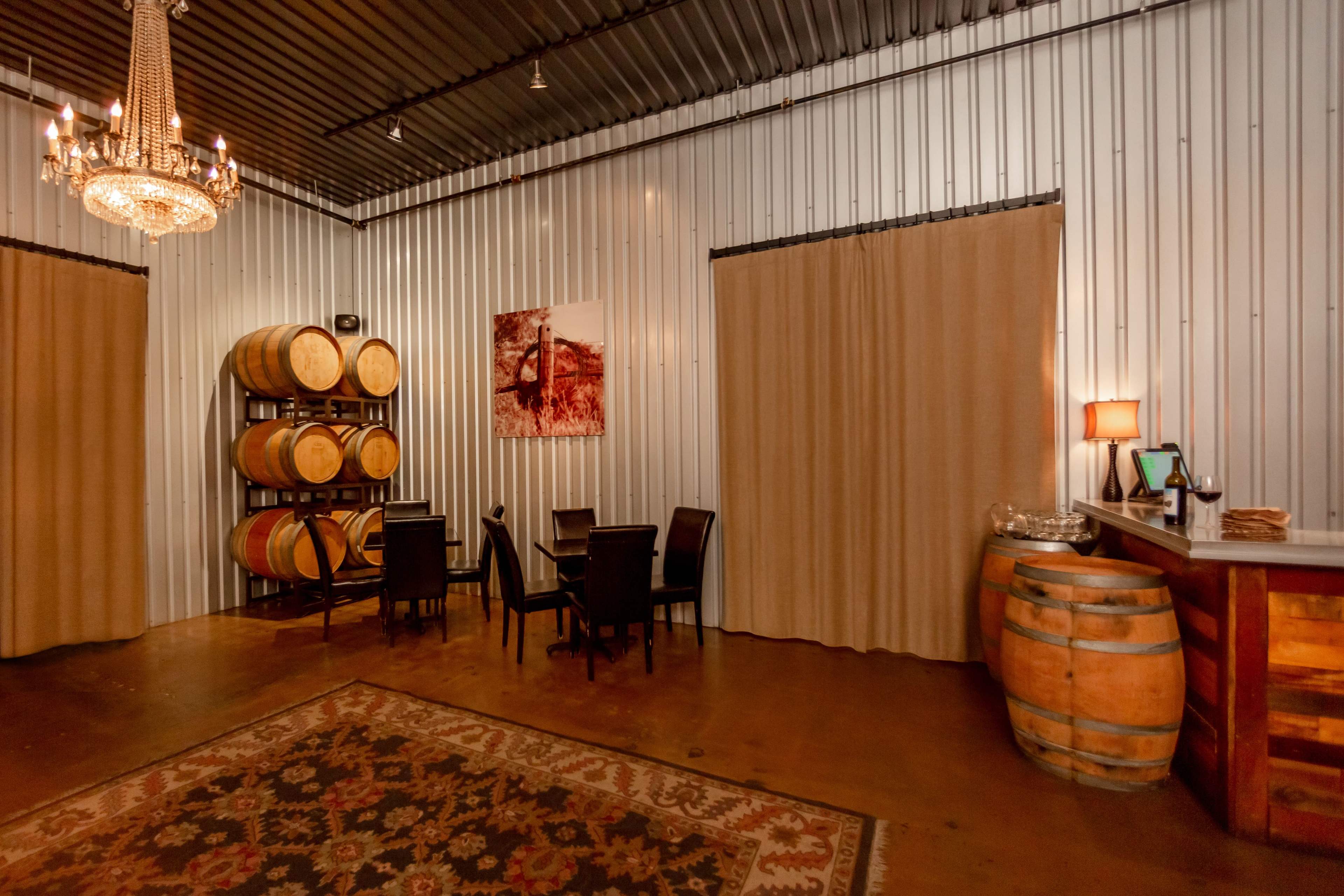 The room features a polished floor, metal walls, a chandelier, wooden wine barrels, and a seating area with dark chairs and a table.
