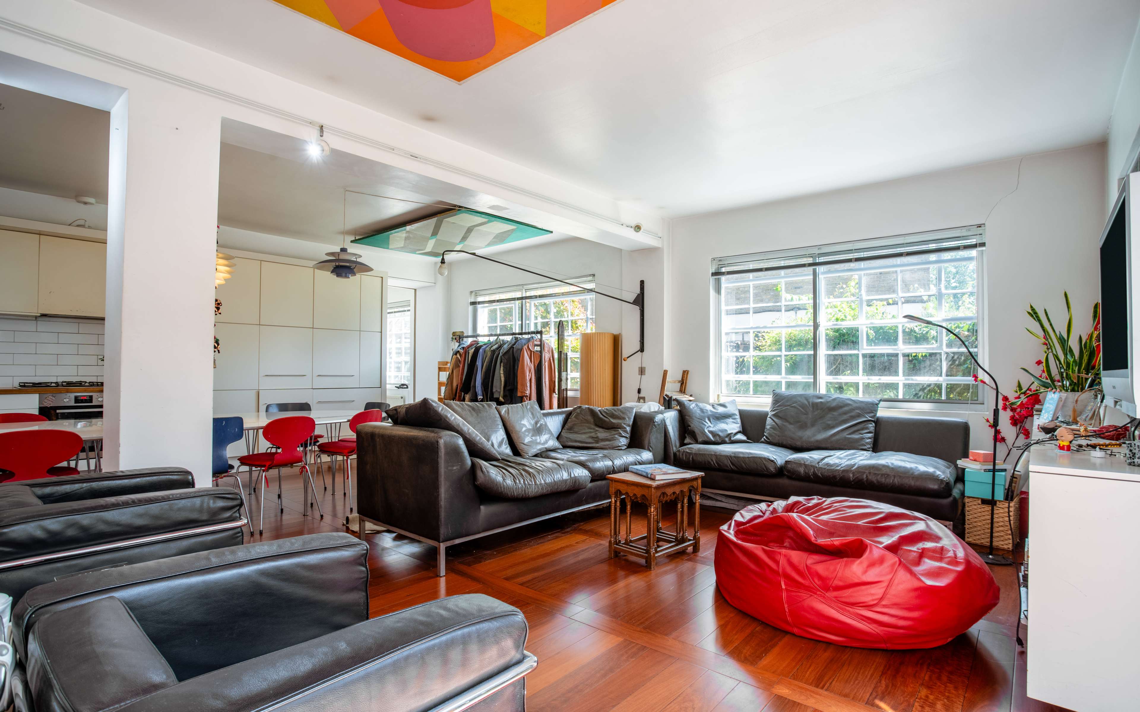 The image shows a modern living room with a large leather sectional, a red bean bag, and dining space in the background featuring a white kitchen and red chairs.