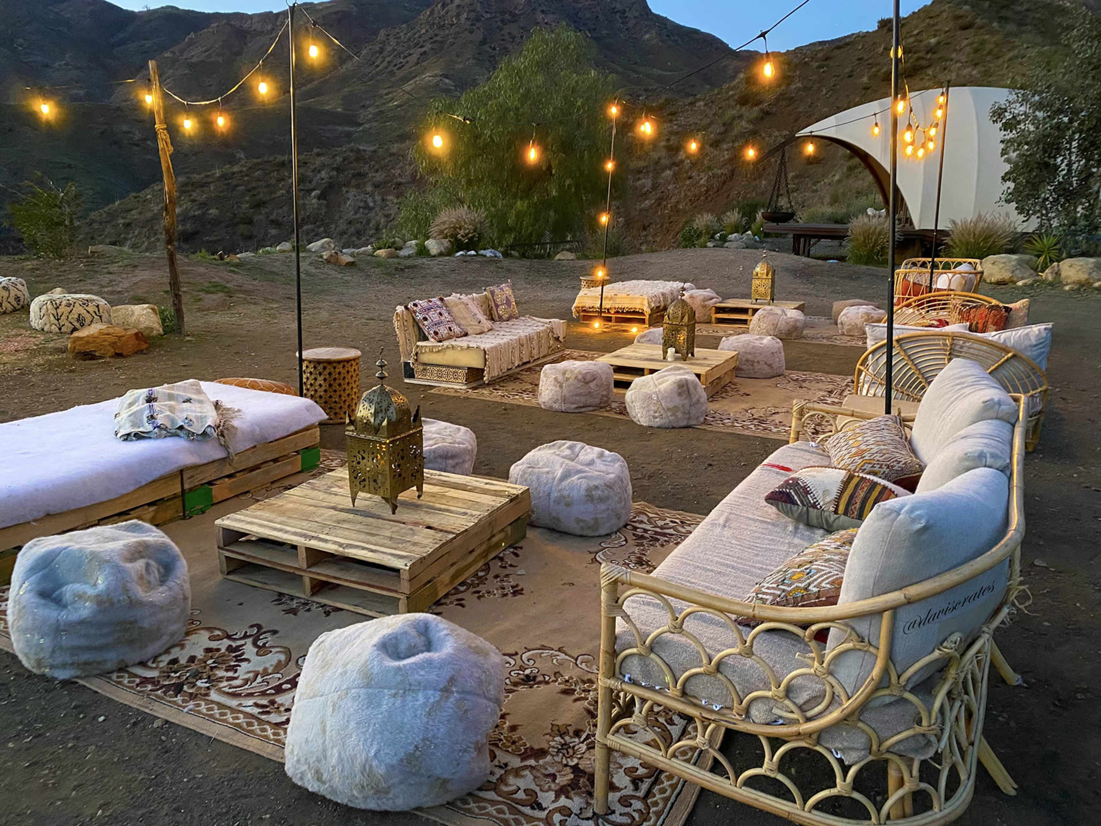 The scene features a rustic outdoor lounge area with string lights, wooden pallet furniture, and soft seating surrounded by mountains.